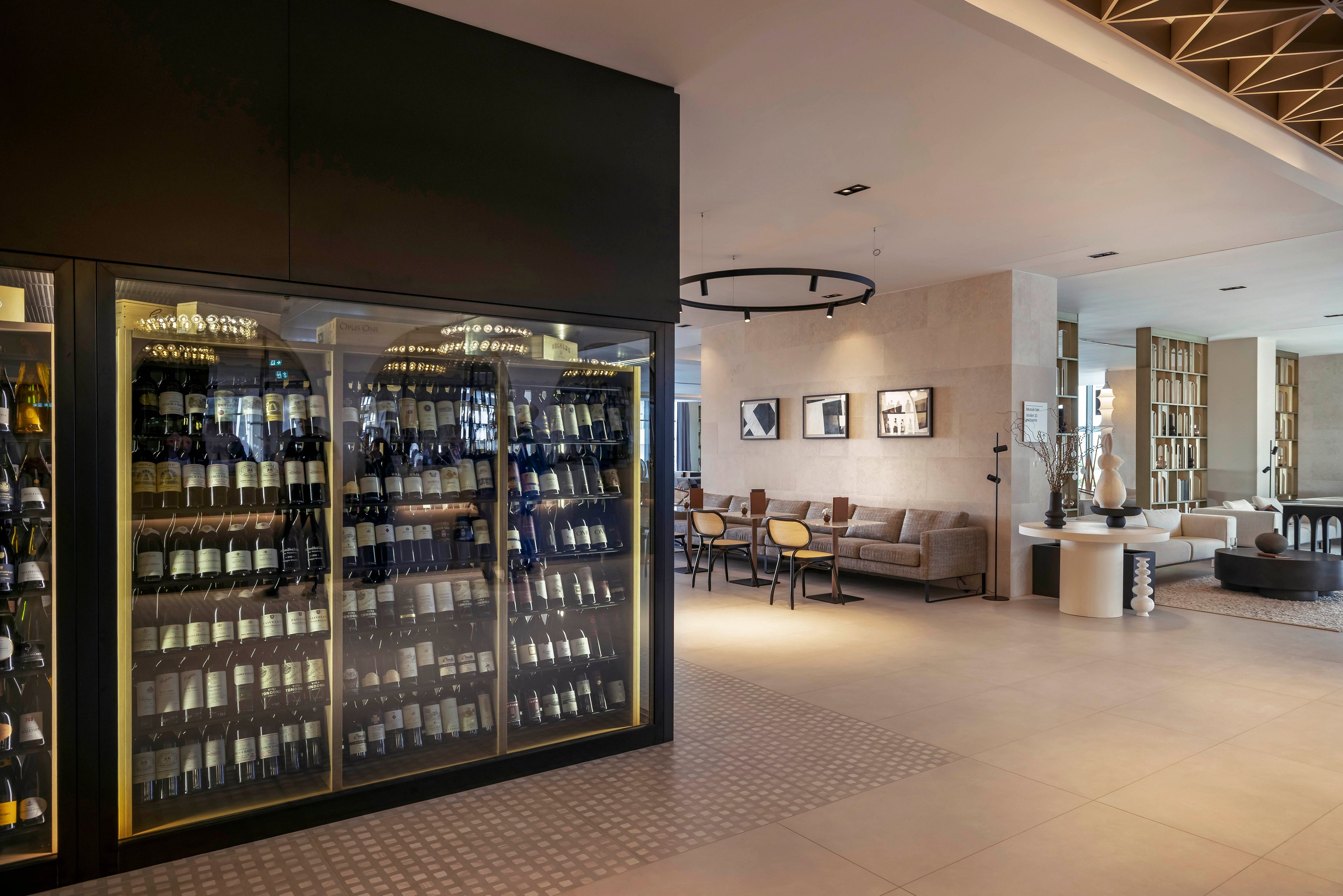 A contemporary hotel lounge features a large glass wine refrigerator filled with assorted bottles. The setting includes a stylish seating area with tables and chairs, neutral tones, and minimalist decor. The space is well-lit with natural and artificial lighting, creating a welcoming and upscale atmosphere. Framed artwork and decorative elements enhance the modern aesthetic.