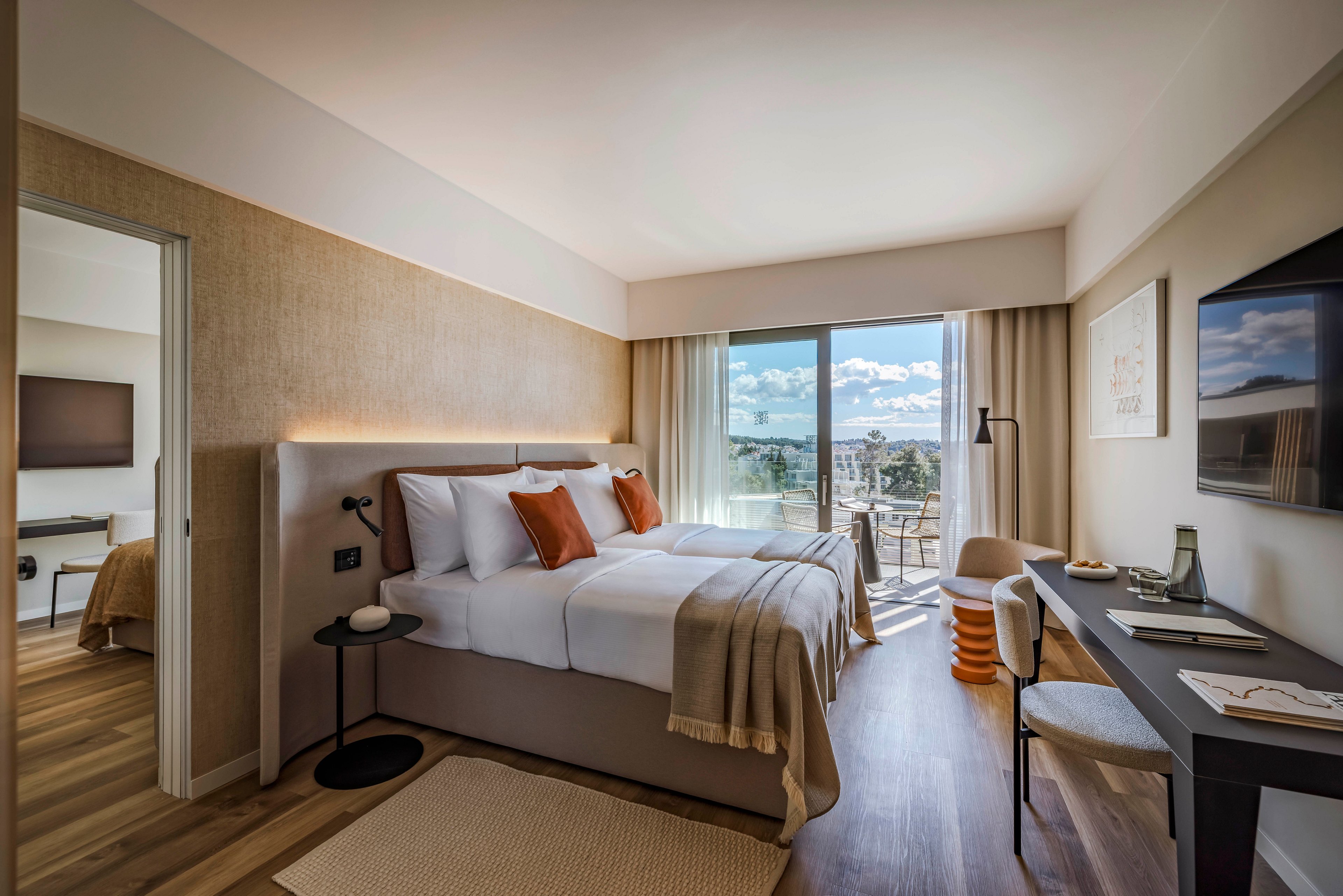 A contemporary hotel room features a neatly made double bed with white linens and brown accent pillows. The space includes a work desk, a wall-mounted television, and a large window leading to a balcony with outdoor seating and scenic views. Warm neutral tones and natural light create a welcoming, relaxed atmosphere. An adjoining room with a television is visible through an open door.