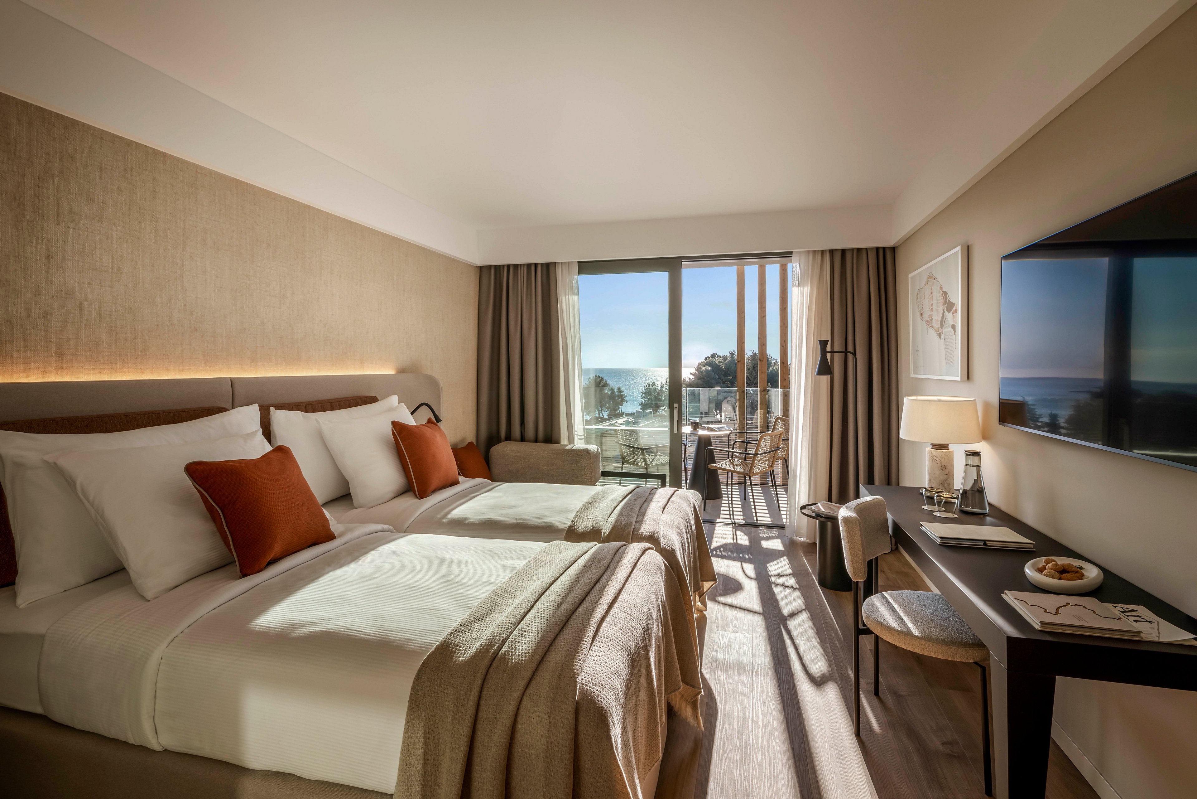 A contemporary hotel room features two neatly made beds with white linens and brown accent pillows. The space is illuminated by natural light streaming through large glass doors that open to a balcony overlooking the sea. The room includes a desk with a lamp, chair, and wall-mounted television, creating a comfortable and inviting atmosphere. Neutral tones and minimalist decor contribute to a calm and relaxing mood.