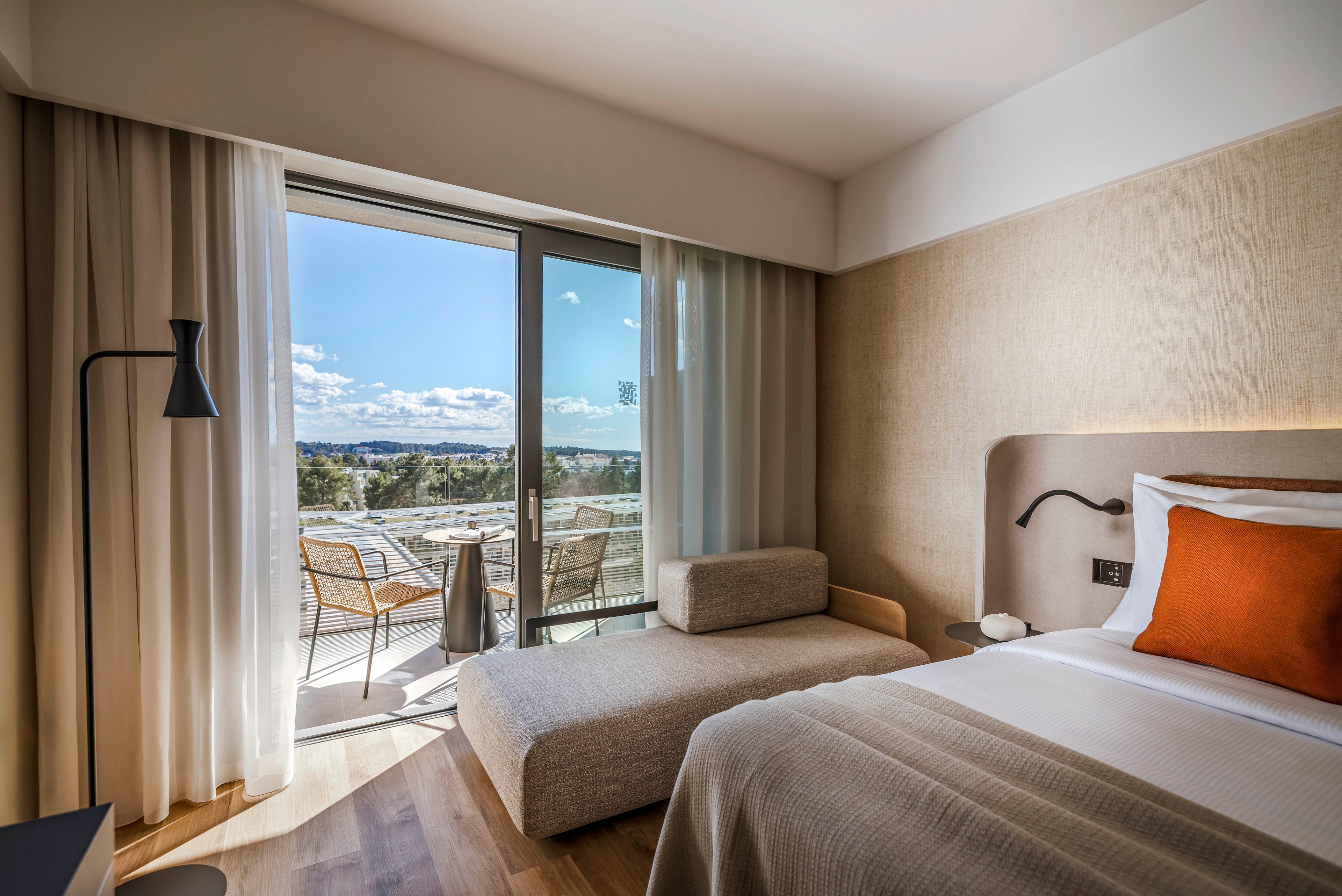 A contemporary hotel room features a neatly made bed with a burnt orange accent pillow and a minimalist chaise lounge. The space opens to a private balcony with two chairs and a table, overlooking a sunny landscape with blue skies and distant greenery. Natural light fills the room, highlighting the neutral color palette and clean, modern design.