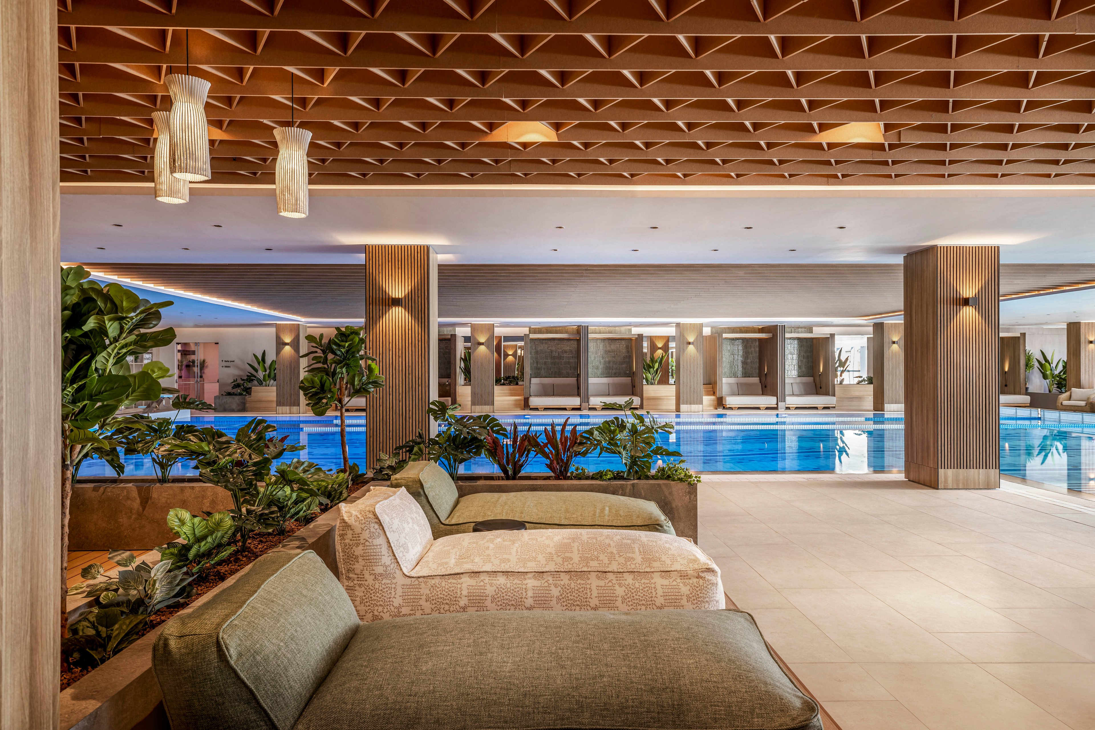 The image features a luxurious indoor swimming pool surrounded by comfortable lounge chairs and lush green plants. The setting is spacious, with a wooden ceiling design and soft ambient lighting, creating a tranquil atmosphere. No people are visible, emphasizing the serene and inviting mood. The palette includes natural wood tones, beige, and blue from the pool water.