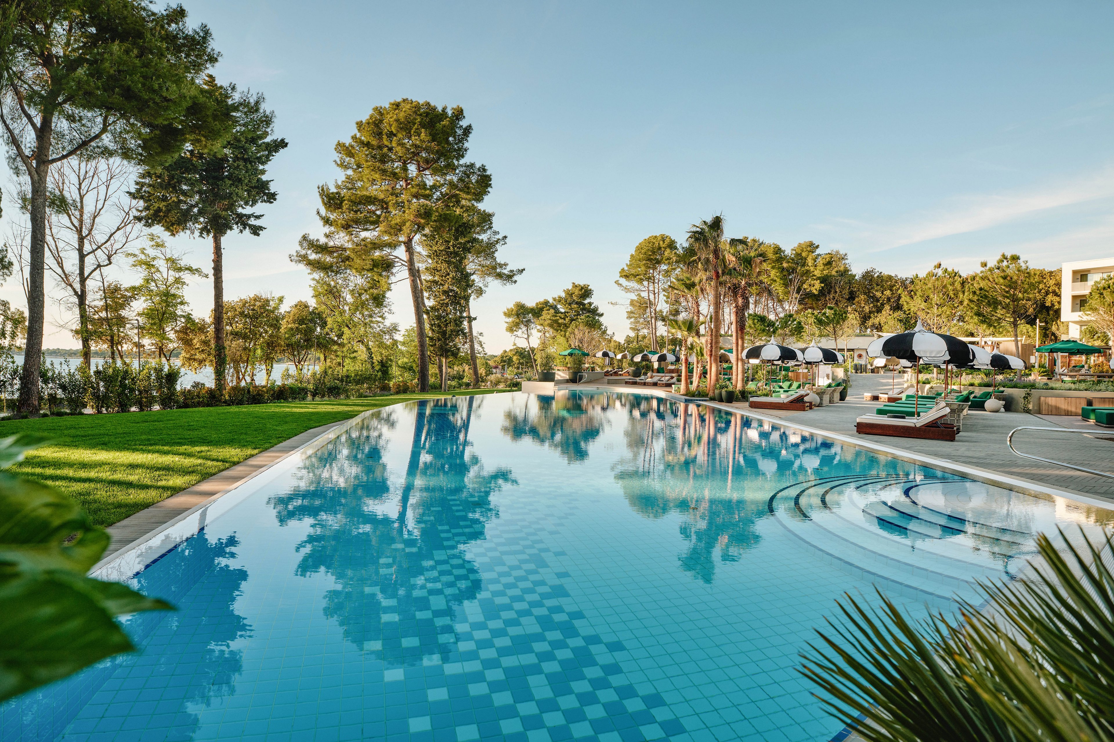 A large, curved swimming pool reflects the surrounding trees and clear morning sky in a tranquil resort setting. Sun loungers and umbrellas line the poolside terrace, suggesting a relaxing vacation atmosphere. The tiled pool surface creates a geometric pattern beneath the water, while landscaped greenery frames the scene. In the background, buildings and distant water hint at a coastal location.