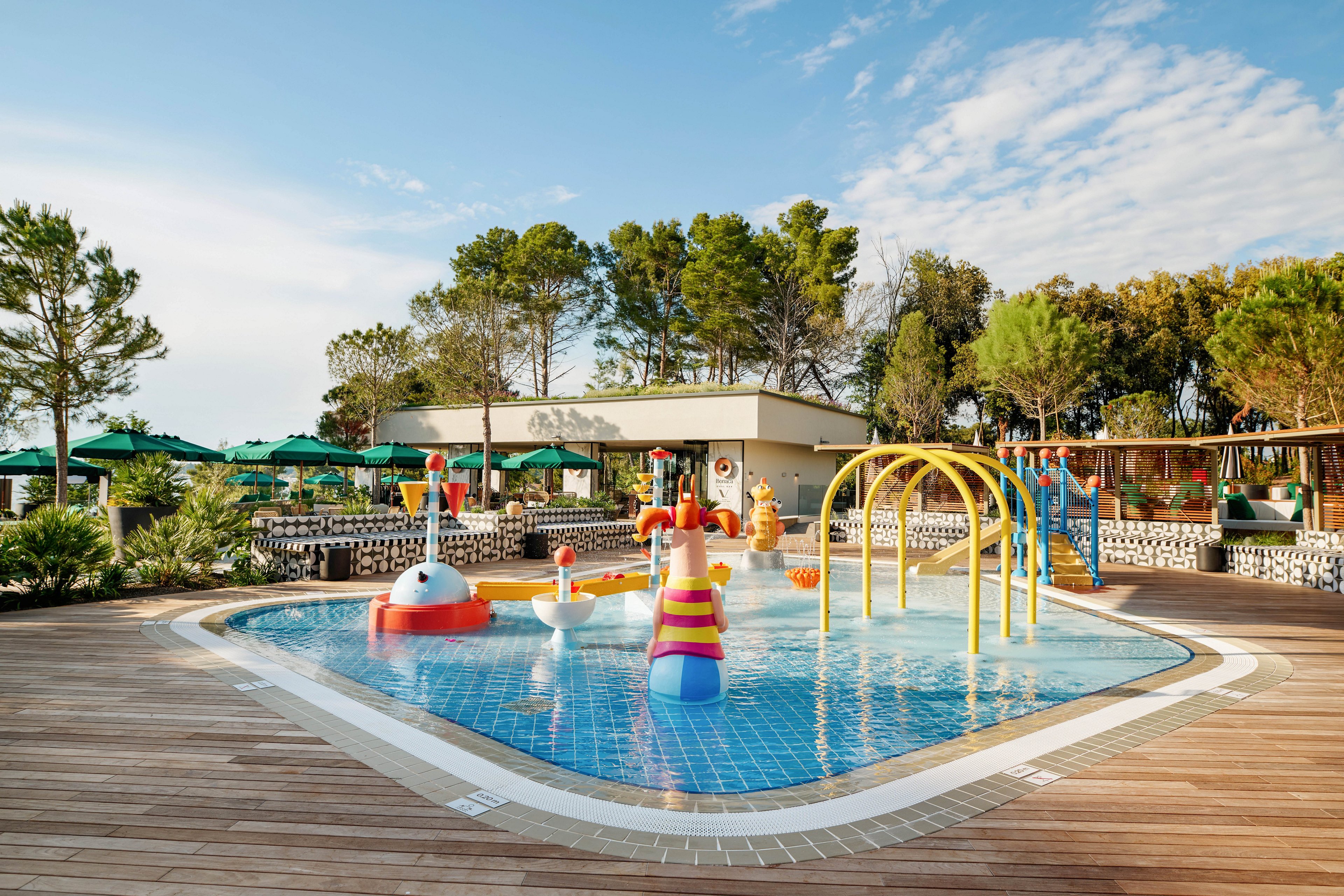 An outdoor splash pad for children features colorful water play structures and fountains set in a shallow tiled pool. Yellow arches spray water over the play area, with whimsical animal and character figures adding a playful touch. Surrounding the pool is a wooden deck with sun loungers and green umbrellas, suggesting a family-friendly resort environment. Tall trees and clear blue sky frame the scene, creating a bright and relaxing holiday atmosphere.