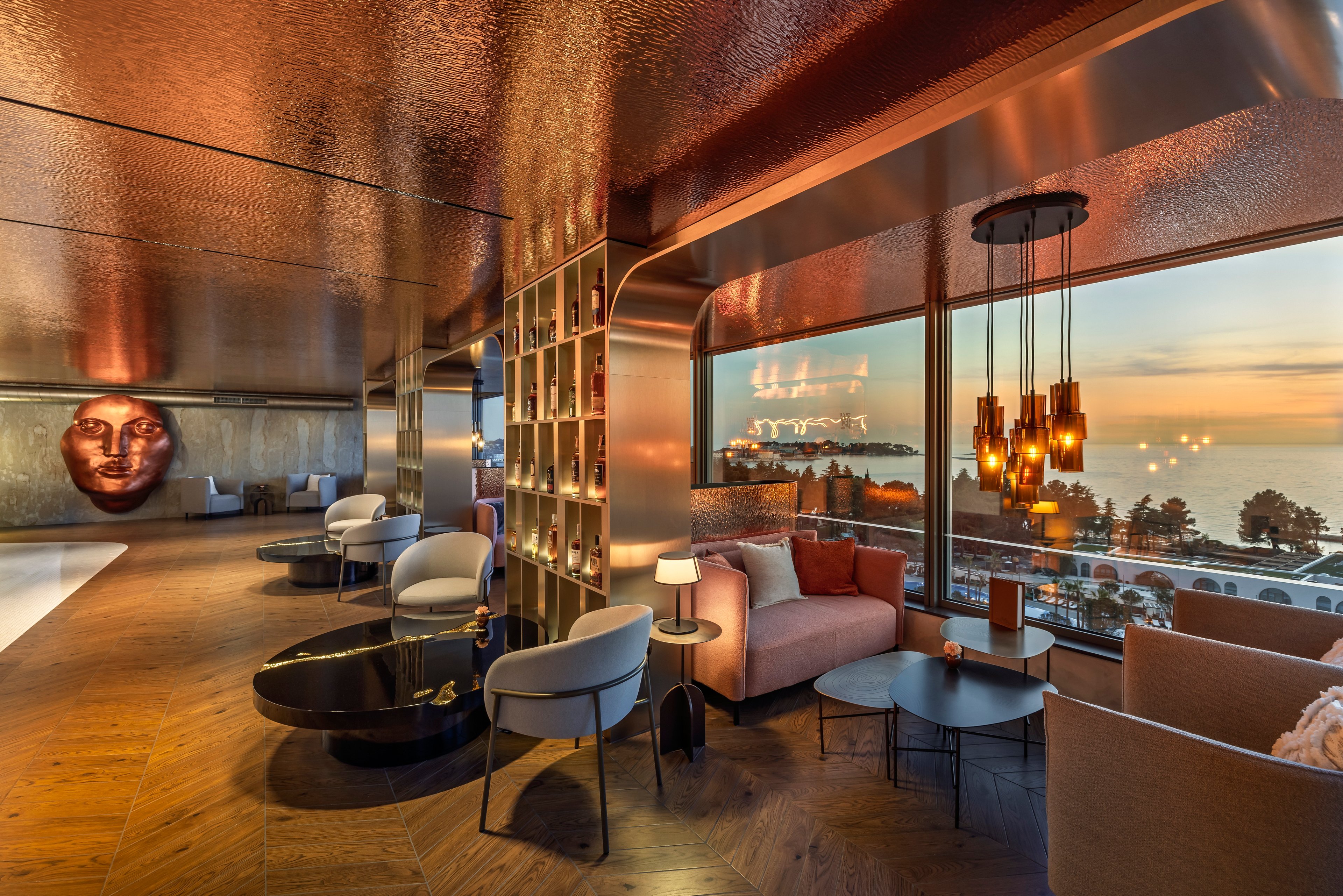 A luxurious hotel lounge features contemporary seating arranged around low black tables and warm accent lighting. Large floor-to-ceiling windows reveal a panoramic view of the sea and coastline at sunset. Copper-toned ceilings and wooden floors create a sophisticated, reflective ambiance. Shelving with decorative objects divides the space, while pendant lights and table lamps add to the cozy, upscale atmosphere.