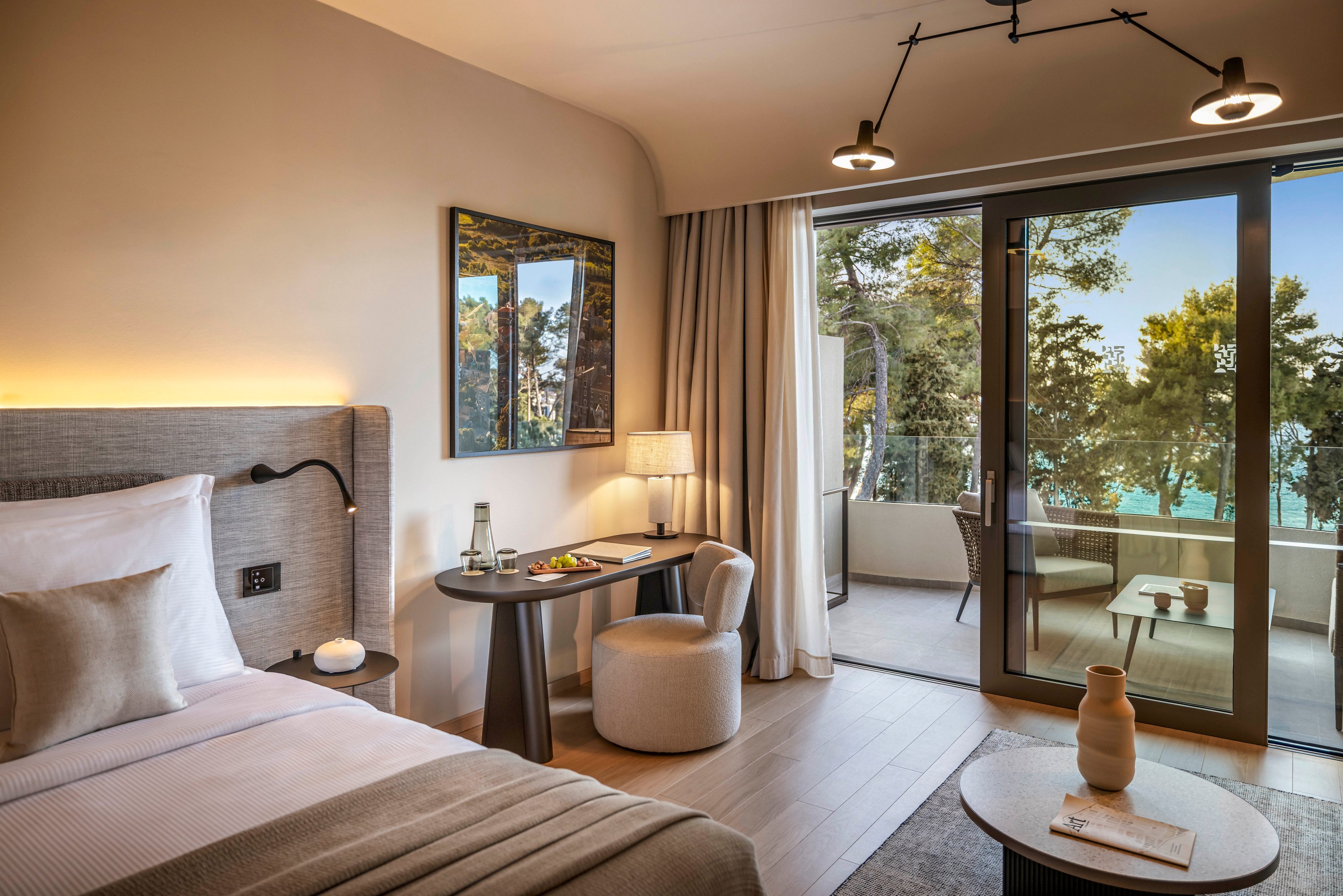 A contemporary hotel room features a neatly made bed, a desk with refreshments, and a cozy seating area. Large glass doors open to a private balcony overlooking lush trees and a glimpse of water. The space is styled in neutral tones with soft lighting, creating a calm and inviting atmosphere. No visible text or numeric values are present in the image.