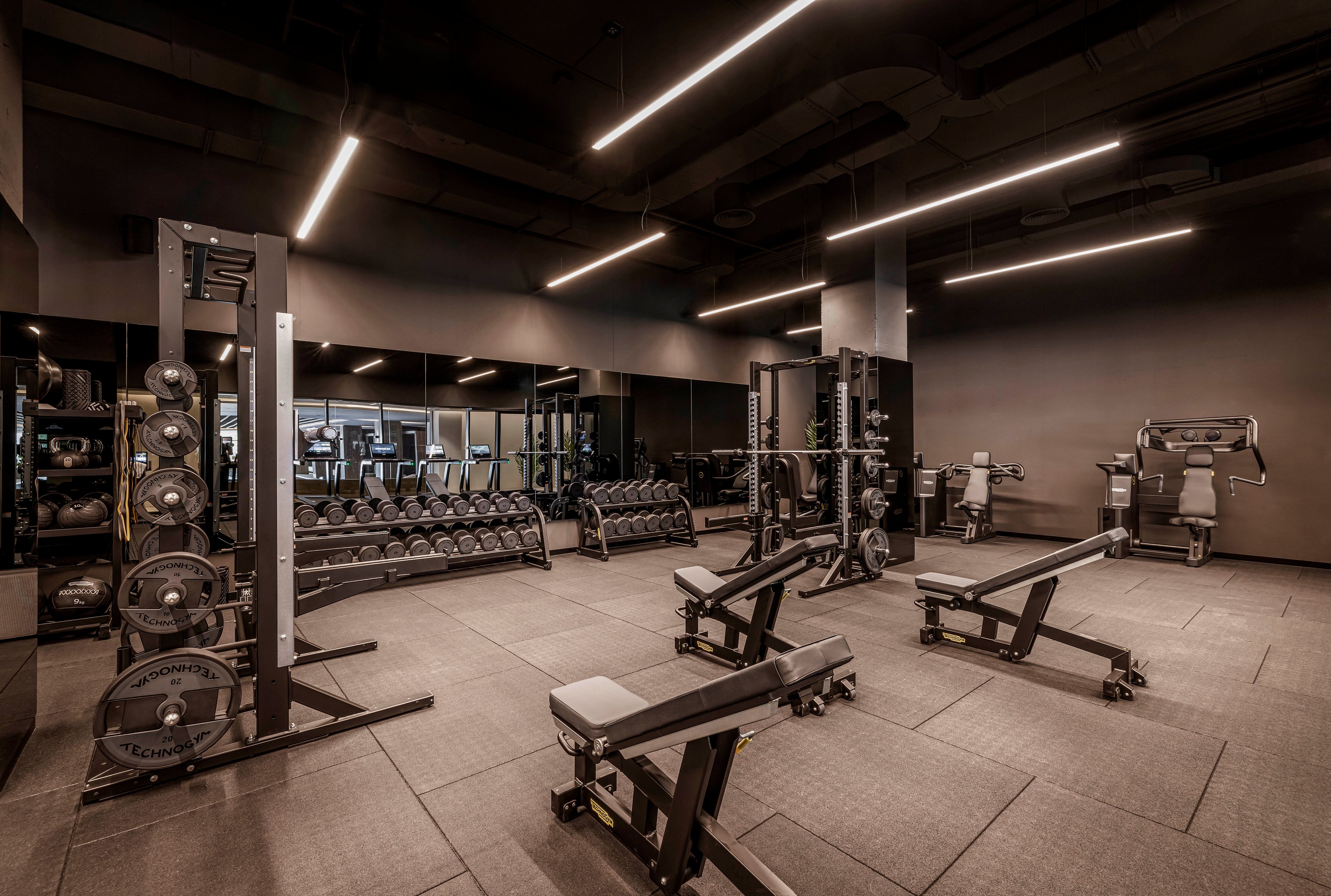 The image shows a spacious, contemporary gym featuring multiple weight benches, dumbbells, and resistance machines. The setting is clean and well-lit with sleek overhead lighting and mirrored walls, creating a professional workout environment. No people are present, emphasizing the equipment and open layout. The color palette is neutral with black and gray tones, contributing to a modern and minimalistic style.
