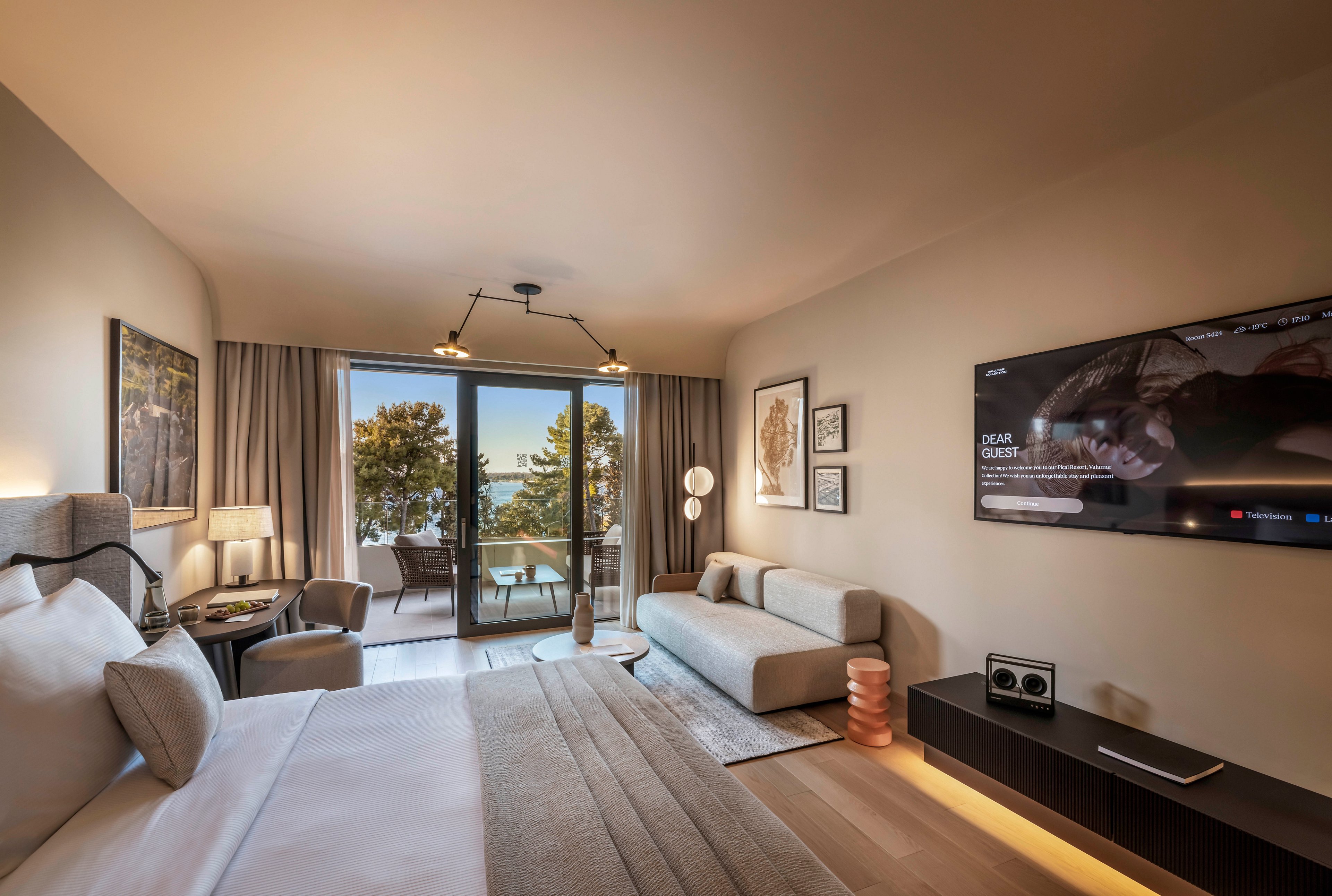 A contemporary hotel room features a large bed, sofa, and workspace, with a wall-mounted smart TV displaying a welcome message. The room opens onto a balcony overlooking trees and outdoor scenery, creating a serene atmosphere. Neutral tones and soft lighting enhance the cozy, inviting mood. Visible text on the TV includes 'DEAR GUEST' and other interface elements.
