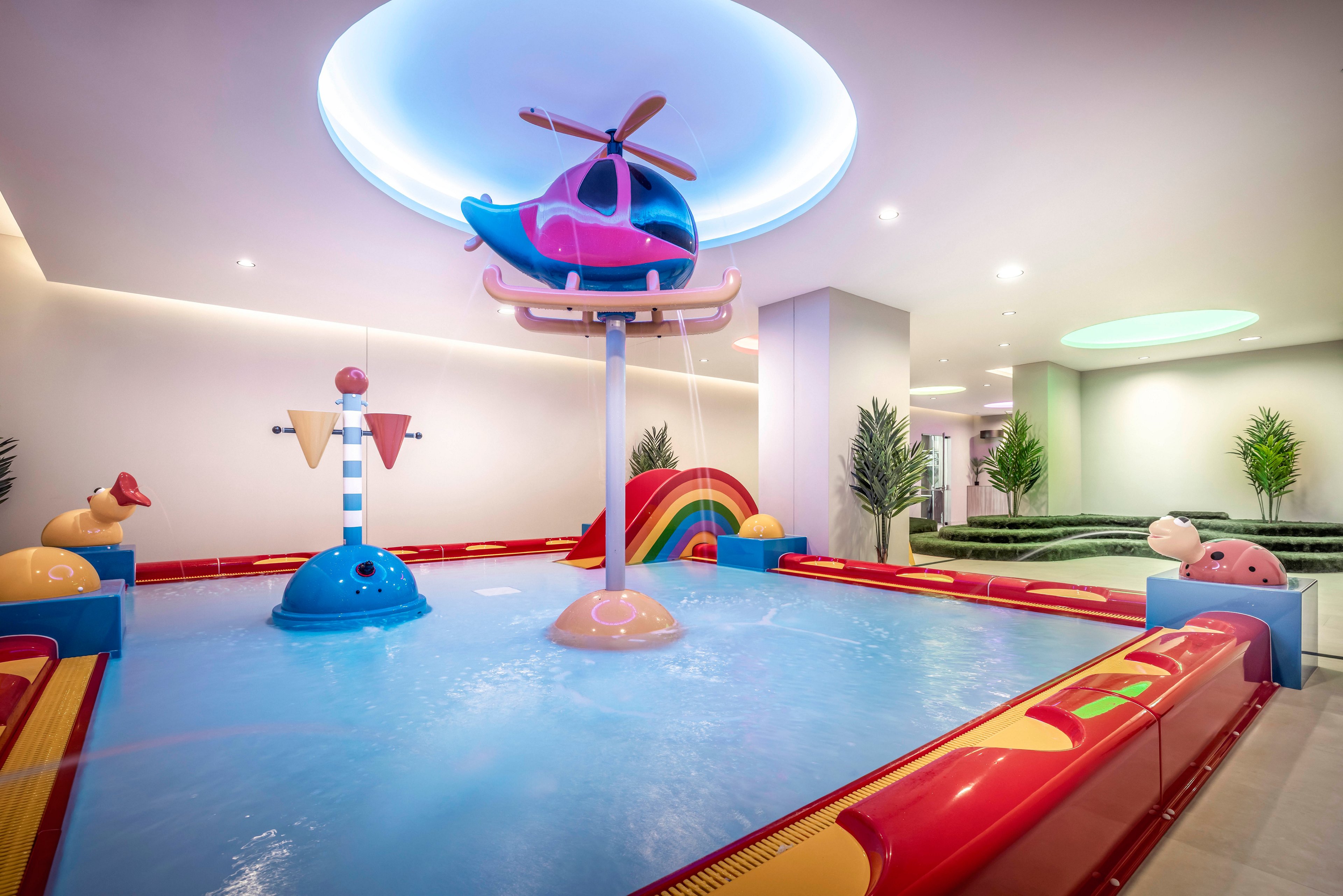 A vibrant indoor pool designed for children features a playful helicopter structure mounted above the water. The setting includes whimsical elements such as a rainbow arch, animal figures, and geometric shapes, all in bright pastel colors. The pool area is illuminated by modern ceiling lights and surrounded by greenery, creating a cheerful and inviting atmosphere. No visible text or numeric values are present in the image.