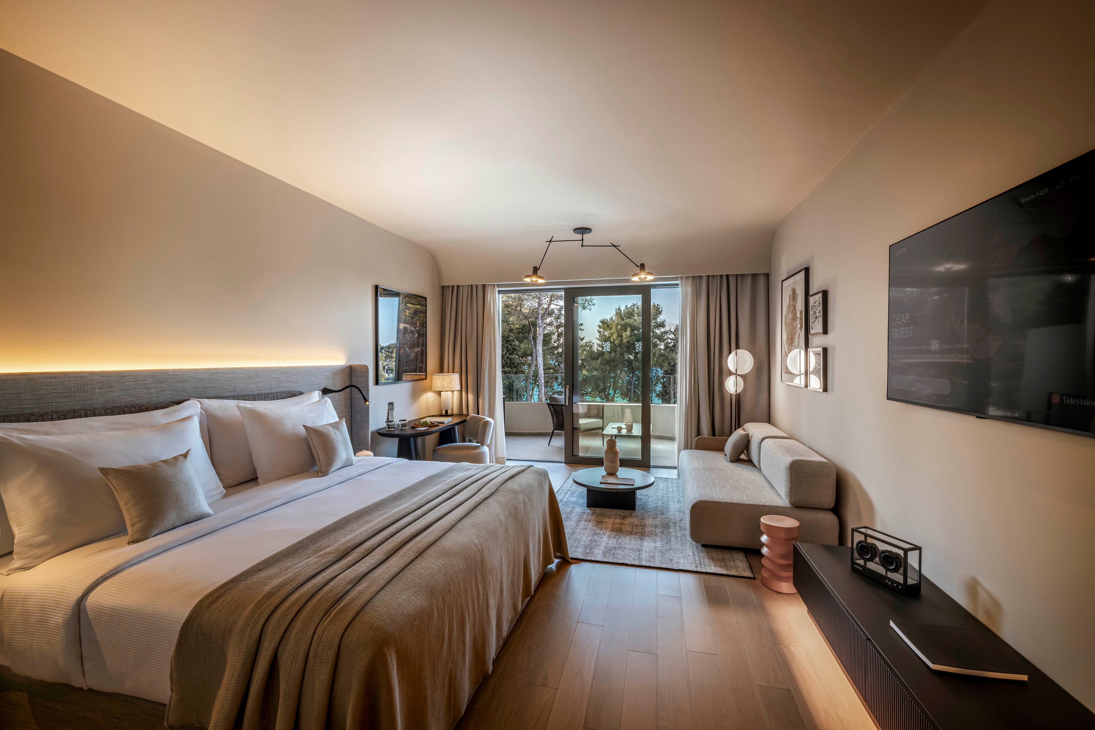 A spacious hotel room features a large bed, cozy seating area, and floor-to-ceiling windows leading to a balcony overlooking trees. The interior is decorated in neutral tones with contemporary furniture and soft lighting. Artwork adorns the walls, and a flat-screen TV is mounted opposite the bed, creating a relaxing and stylish atmosphere.
