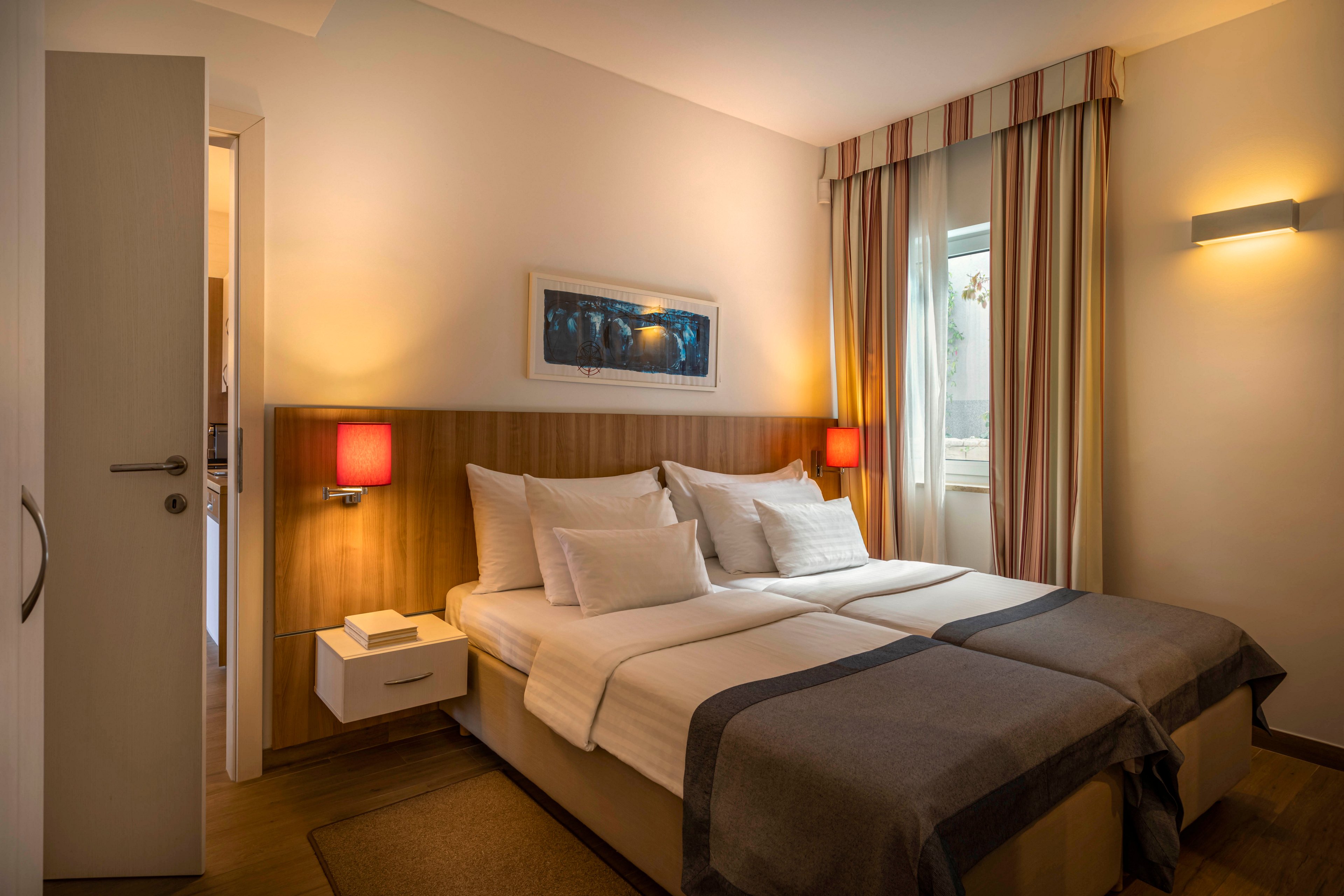 A contemporary hotel room features a neatly made double bed with crisp white linens and multiple pillows. The setting includes warm lighting from two red bedside lamps, a wooden headboard, and a window with striped curtains. The room is decorated with a wall art piece above the bed and a small nightstand. The overall mood is cozy and inviting, with neutral and earthy tones.