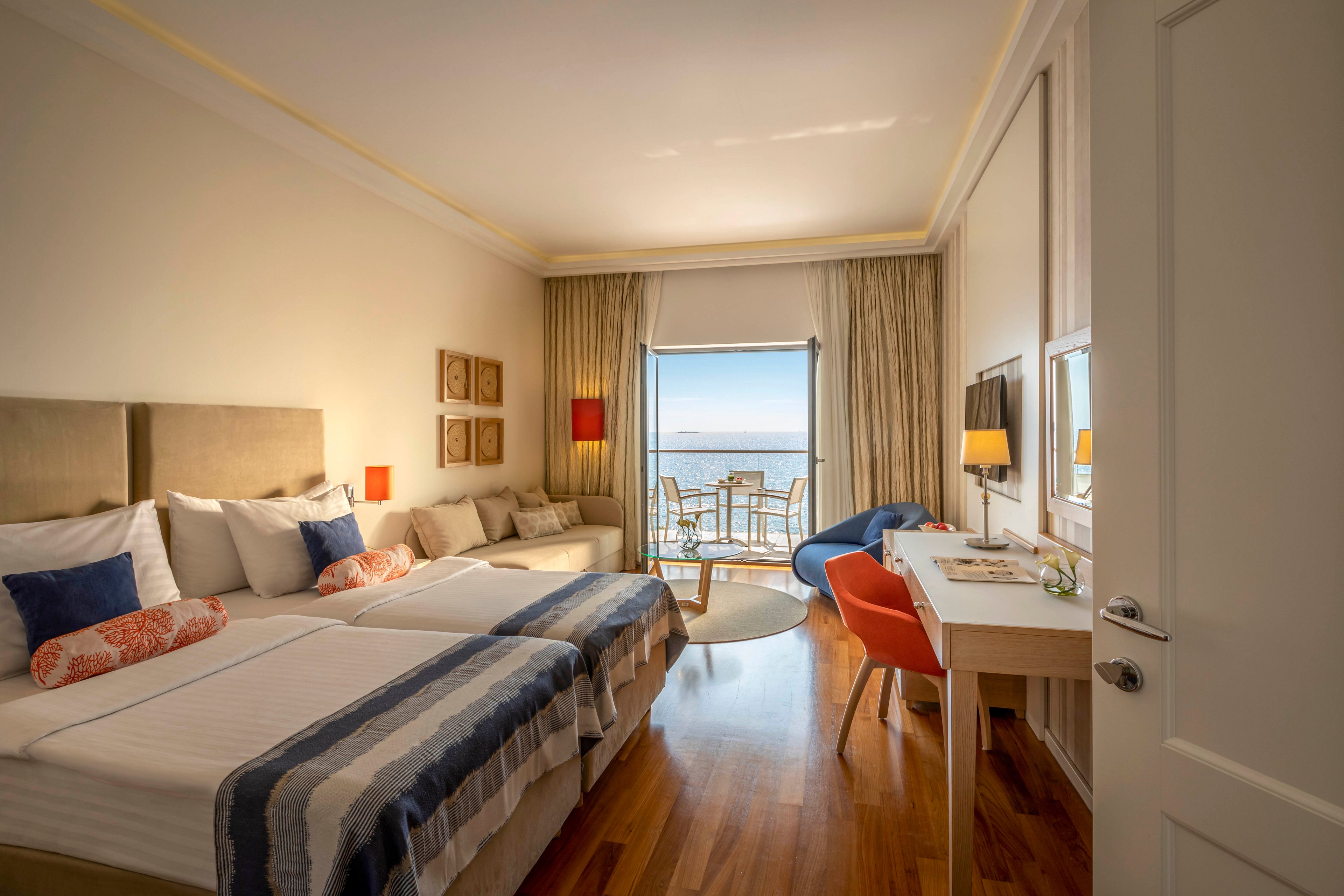 A spacious hotel room features two neatly made beds with patterned throws and accent pillows. The room includes a desk, television, and seating area, all illuminated by natural light from a balcony overlooking the ocean. Warm neutral tones and wooden flooring create a cozy, inviting atmosphere, with pops of orange and blue adding visual interest. The balcony offers outdoor seating and a scenic water view.