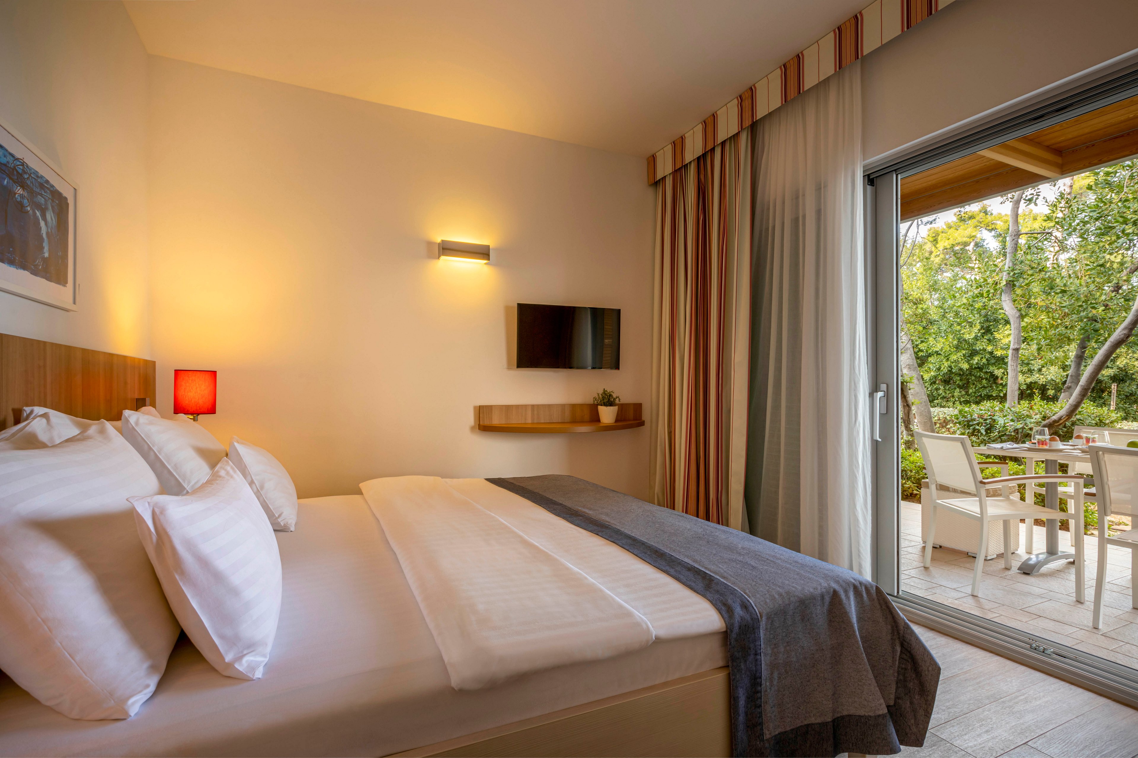 A contemporary hotel room features a neatly made bed with white linens and a dark throw blanket. The room includes a wall-mounted television, a small shelf, and warm lighting from a red lamp. Large sliding glass doors open to a patio with outdoor seating, overlooking a lush green garden. The setting is calm and inviting, with neutral tones and natural light.