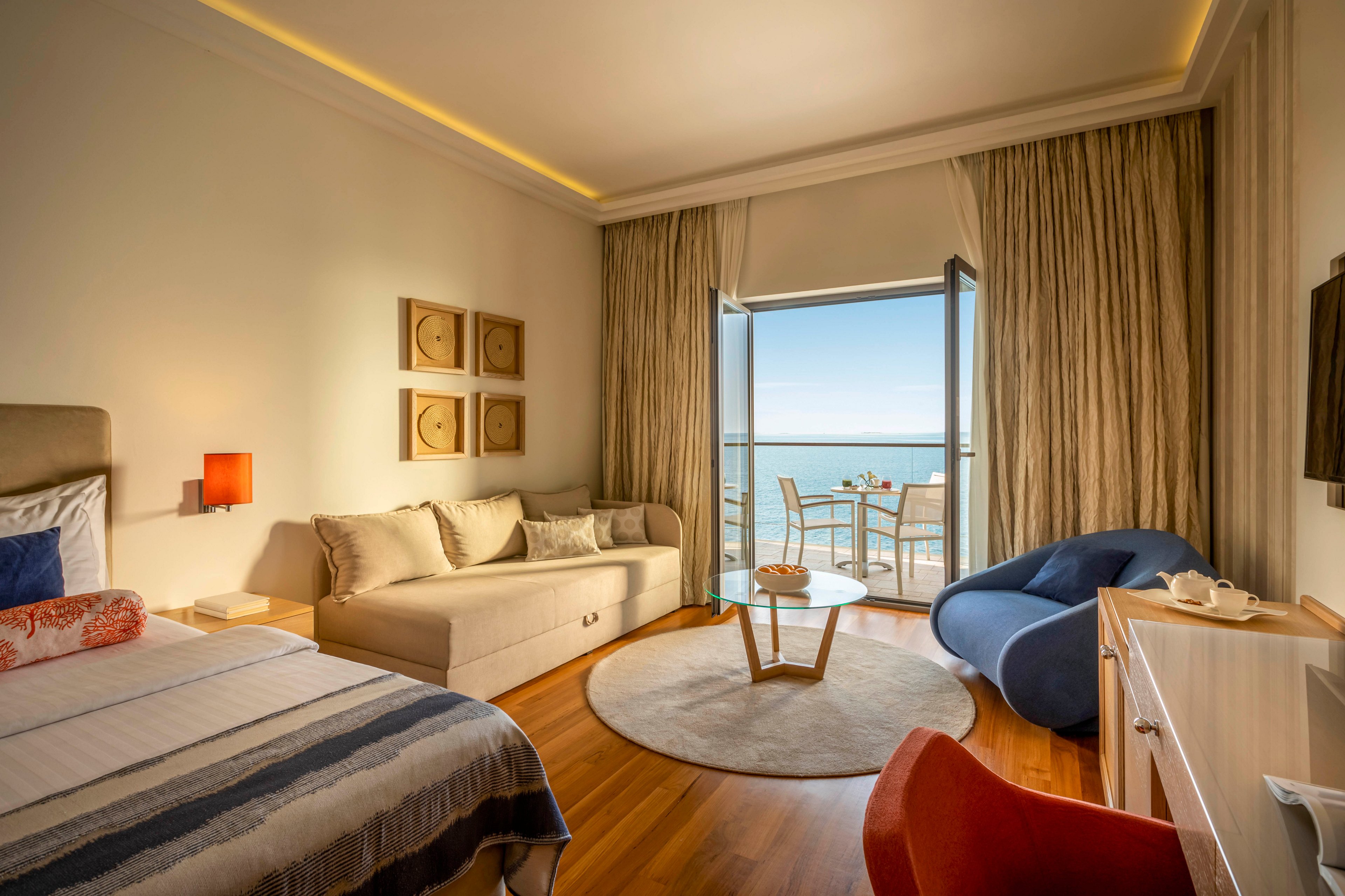 A contemporary hotel room features a cozy bed, sofa, and stylish seating area. The space opens onto a balcony with a table and chairs overlooking the ocean, creating a serene atmosphere. Warm wood flooring, neutral tones, and soft lighting enhance the inviting mood. No visible text or numeric values are present in the image.