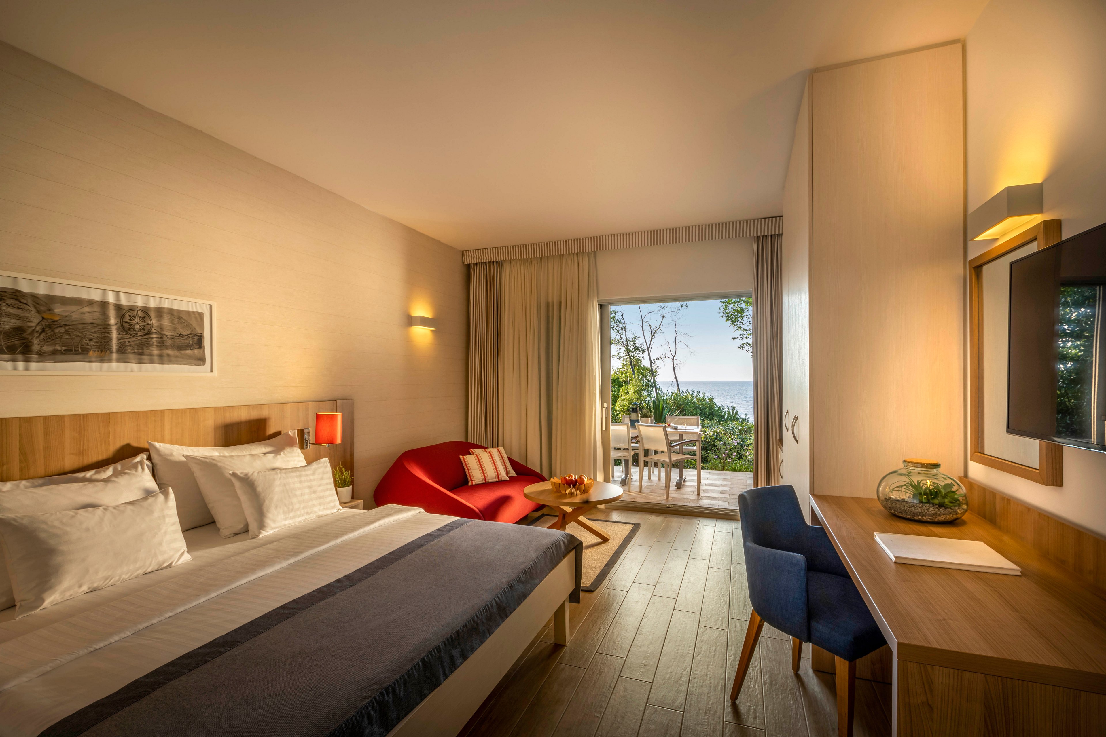 A contemporary hotel room features a large bed, a red sofa, and a work desk with a chair. The room opens to a balcony with outdoor seating, overlooking a scenic view of the sea and greenery. Warm lighting and neutral tones create a cozy, inviting atmosphere, while natural light streams in through the glass doors.