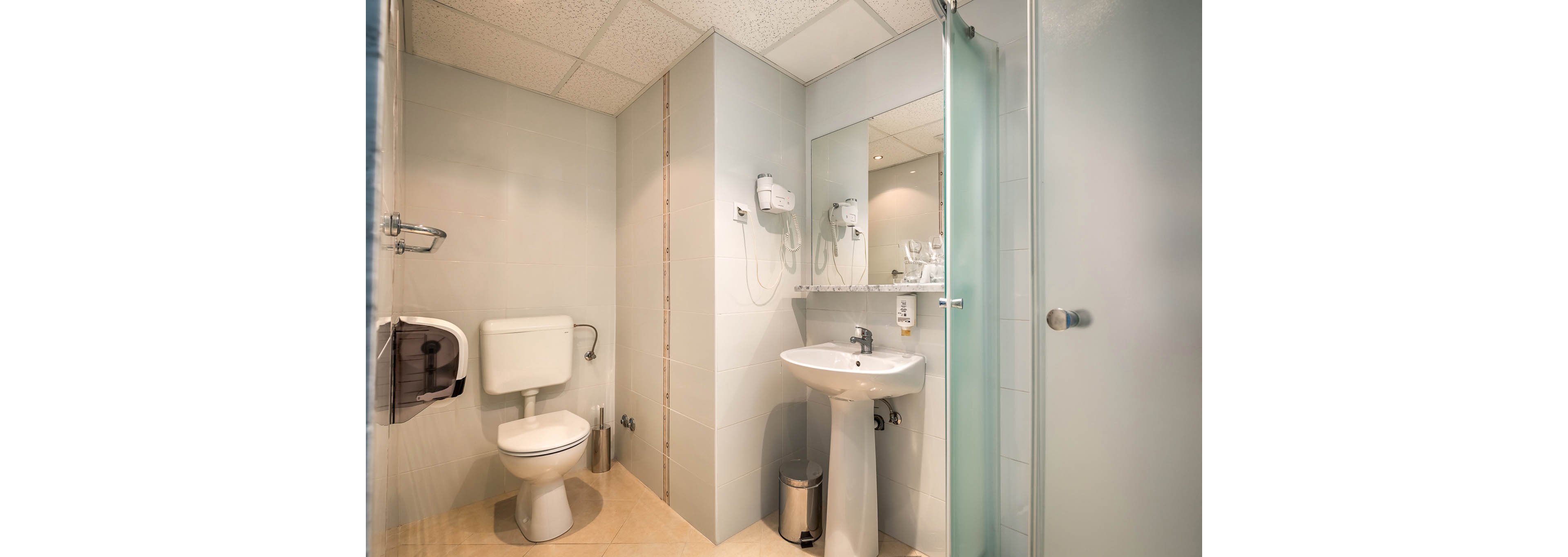 A clean, modern bathroom features a white toilet, pedestal sink, and a glass shower enclosure. The setting includes a large wall mirror, mounted hair dryer, and a stainless steel trash bin. Neutral tiles and soft lighting create a bright, inviting atmosphere. No visible text or numbers are present in the image.