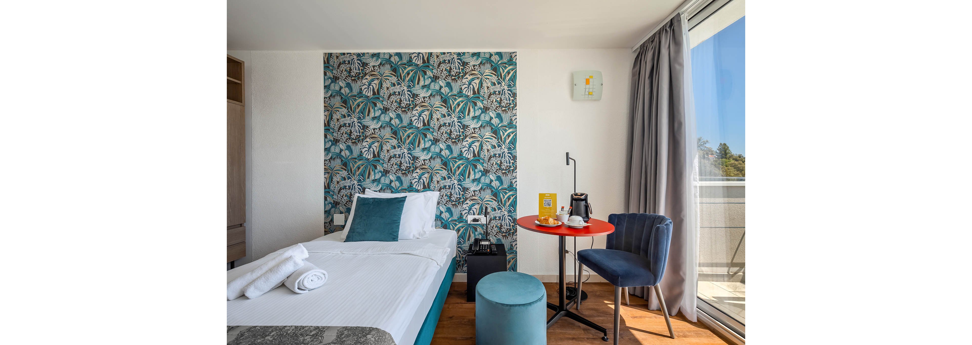 A contemporary hotel room features a single bed with white linens and teal accents, set against a bold tropical-patterned accent wall. The space includes a small round table with a coffee maker, cups, and a breakfast tray, as well as a plush blue chair and ottoman. Natural light streams in from a large window with gray curtains, revealing a balcony outside. The room is styled in a modern, inviting manner with a mix of teal, blue, and neutral tones. A rolled towel is placed on the bed, adding a welcoming touch.