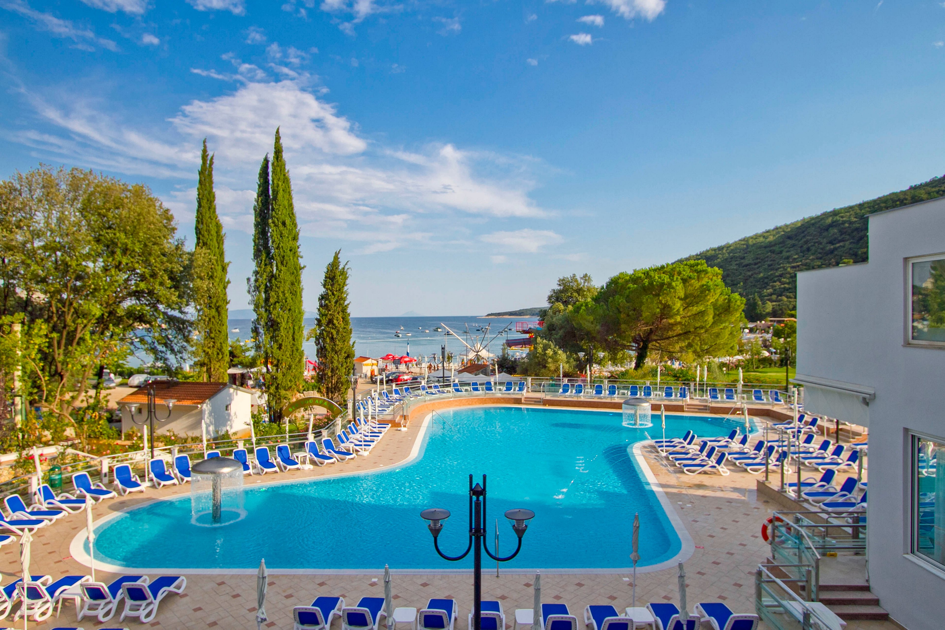 A large outdoor swimming pool is surrounded by numerous blue and white lounge chairs, set in a resort environment. The pool area is bordered by lush greenery and tall trees, with a view of the sea and distant hills in the background. The sky is partly cloudy, and the overall mood is tranquil and inviting, ideal for relaxation. Modern white buildings and decorative lamp posts add to the resort ambiance.
