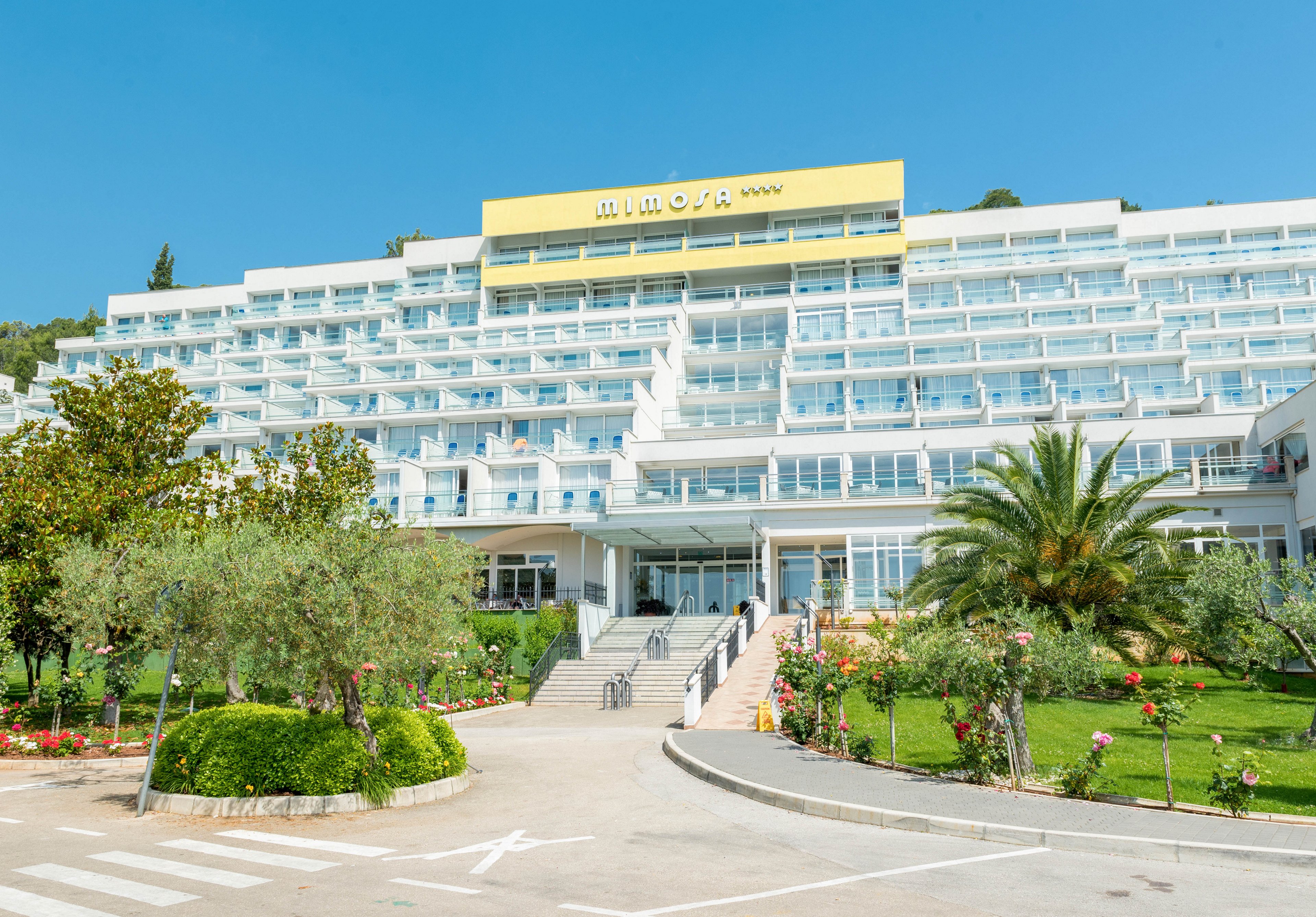 A large, multi-story hotel building is shown under a clear blue sky, with a prominent yellow sign reading 'IMPERIAL' and four stars. The hotel features numerous balconies and glass windows, surrounded by well-maintained gardens with palm trees, flowering bushes, and trimmed greenery. The setting is bright and inviting, evoking a relaxed, vacation atmosphere.