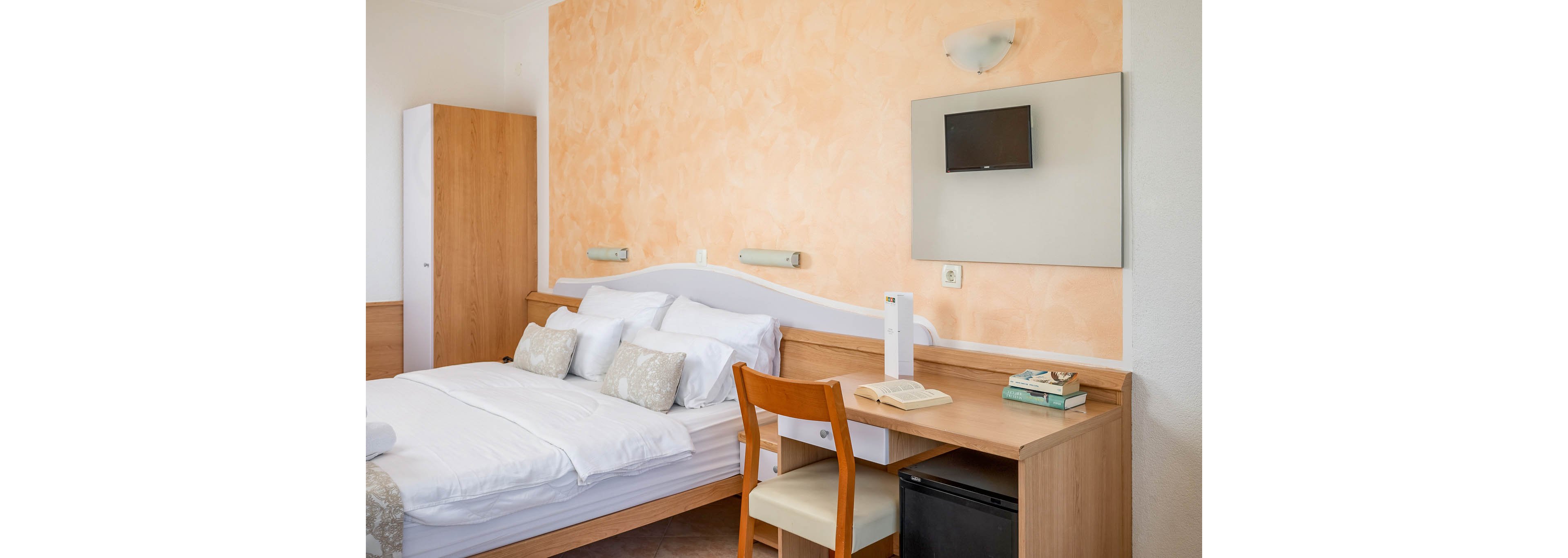 A neatly arranged hotel room features a double bed with white linens and decorative pillows. The space includes a wooden desk with a chair, books, and an open notebook, set against a peach-toned textured wall. A small wall-mounted television and a wardrobe are also visible, creating a cozy and functional atmosphere. The lighting is soft and natural, enhancing the inviting mood.