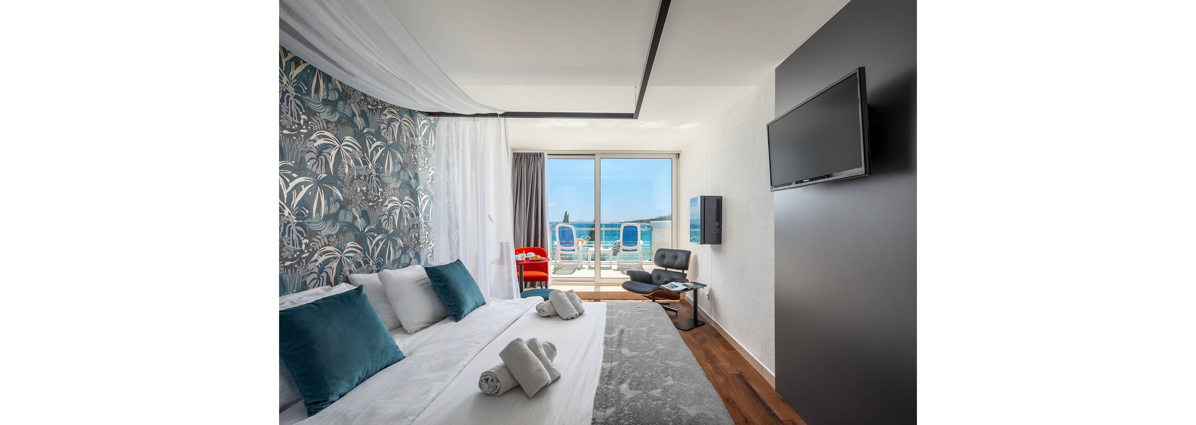 A contemporary hotel room features a large bed with teal accent pillows and rolled towels, set against a decorative botanical wallpaper. The space opens to a balcony with two blue lounge chairs overlooking the ocean, creating a relaxing atmosphere. The room includes a wall-mounted flat-screen TV, a sleek black chair, and a small table, with natural light streaming in through glass doors. The overall palette is cool and neutral, emphasizing comfort and tranquility.