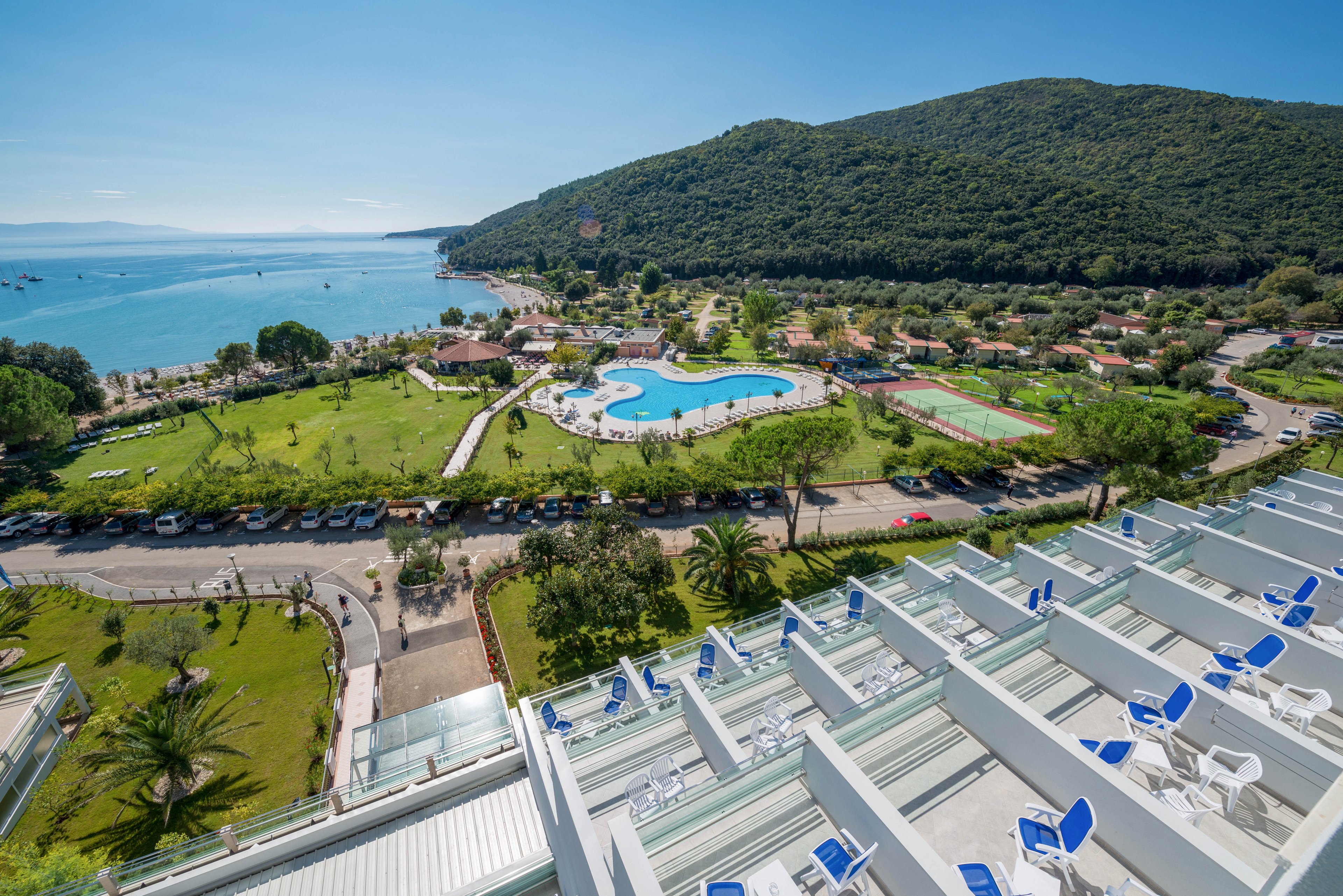 A series of modern white balconies with blue chairs face a large outdoor swimming pool and tennis courts. The resort is set in a lush green landscape near the coastline, with a mountain rising in the background. The scene is bright and sunny, evoking a relaxed vacation atmosphere. No visible text or numbers are present in the image.