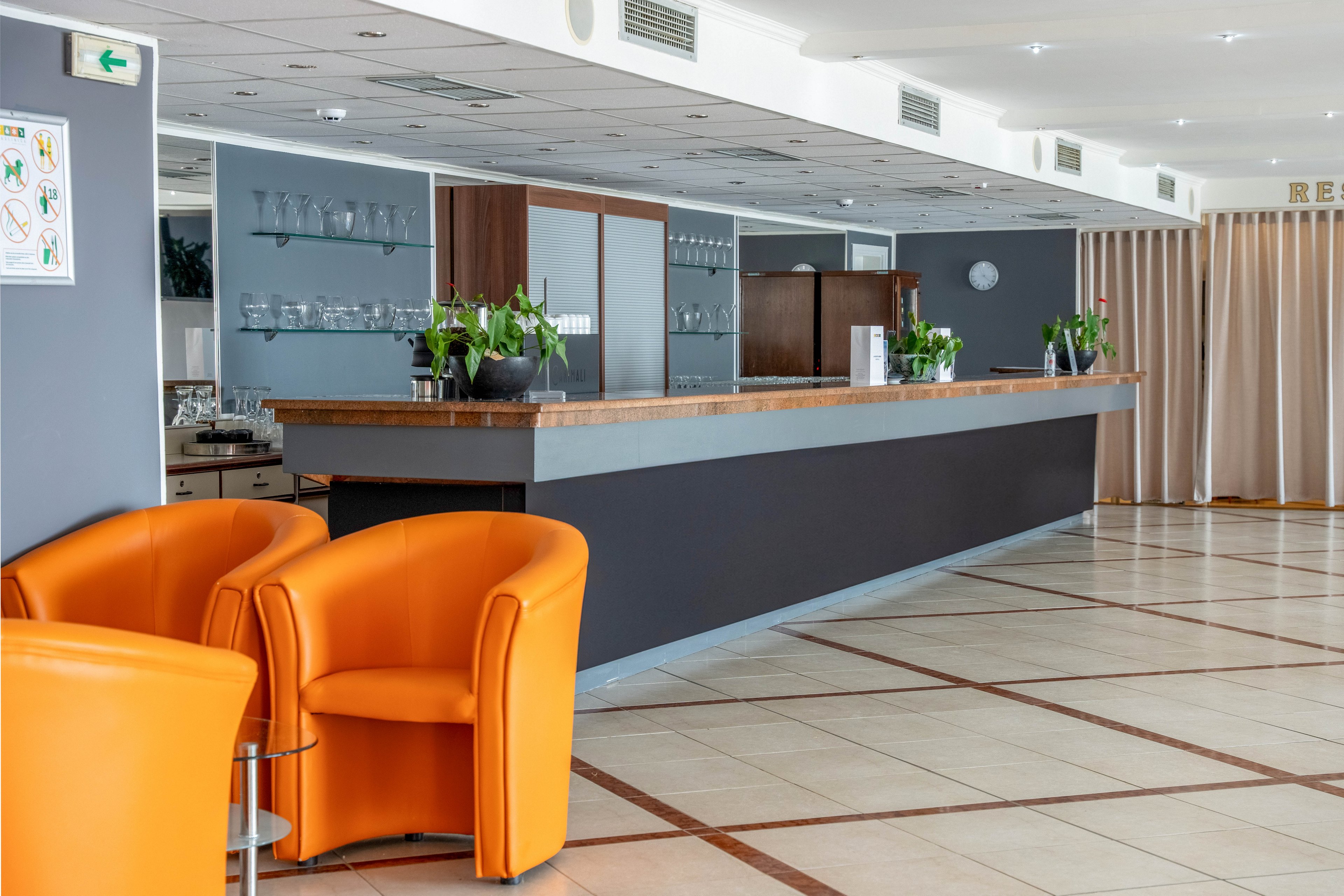 A contemporary hotel lobby features a sleek bar counter with wooden accents and several potted plants. Two bright orange armchairs are positioned in the foreground on a tiled floor with geometric patterns. The setting is well-lit, with glassware visible behind the bar and neutral-colored walls creating a clean, inviting atmosphere.