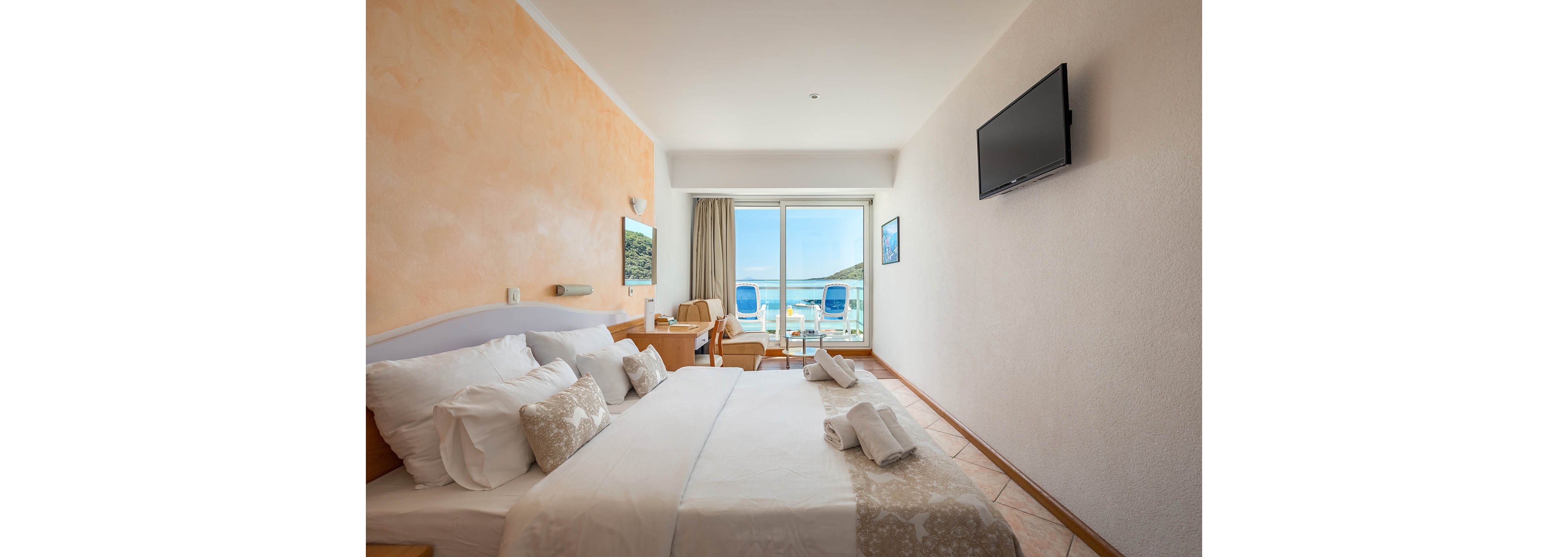 A neatly arranged hotel room features a large bed with white linens and decorative pillows. The room opens to a balcony with two blue chairs overlooking the ocean, creating a relaxing coastal atmosphere. A wall-mounted television and a small desk area are visible, with soft natural light filling the space. The overall palette is neutral with touches of beige and blue.