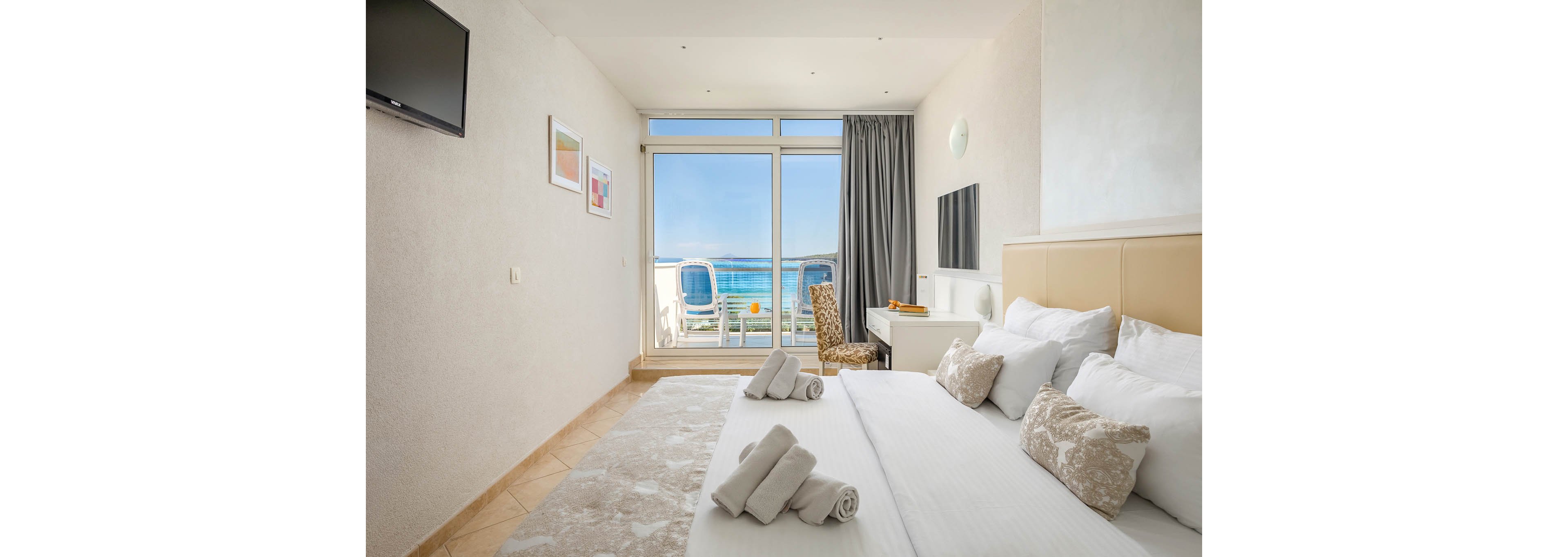 A modern hotel room features a large bed with multiple pillows and rolled towels, facing a wall-mounted television. The room opens to a balcony with two chairs and a stunning view of the ocean. Neutral tones and natural light create a calm, inviting atmosphere, with a desk and chair positioned near the window. Framed artwork decorates the walls, and a curtain partially covers the glass door. No visible text or numbers are present in the image.