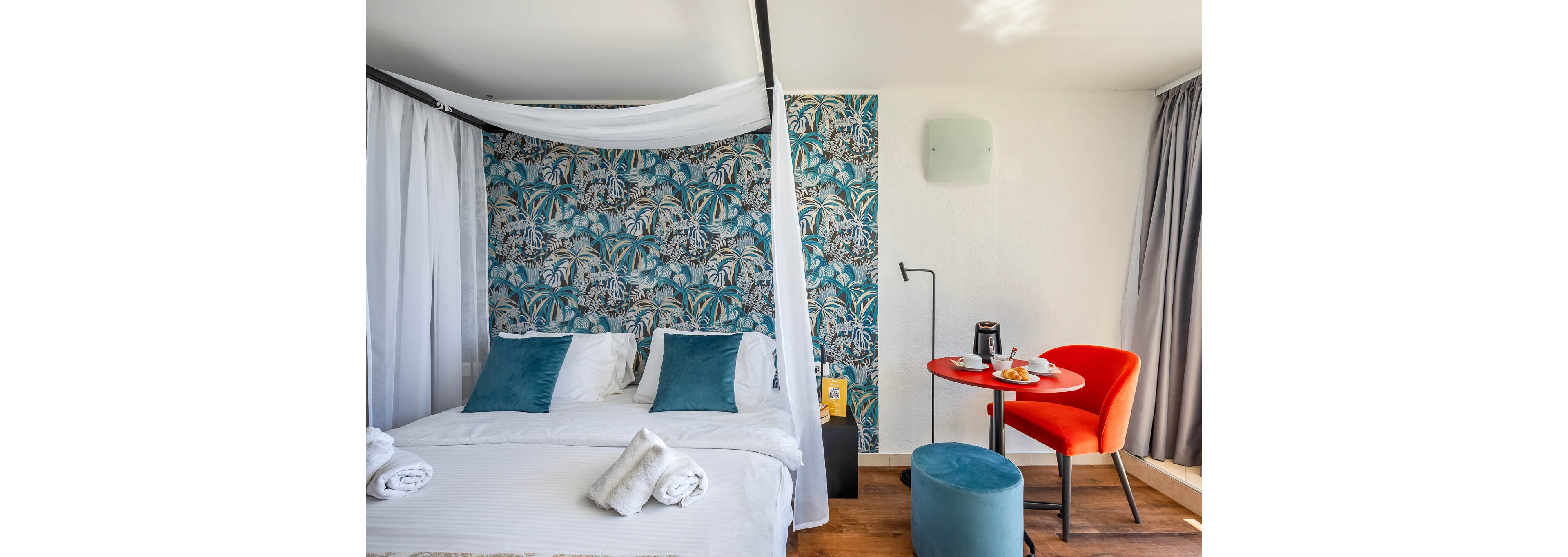 A stylish hotel room features a white canopy bed with teal accent pillows and a tropical-patterned wallpaper. A small red table is set for breakfast with coffee, juice, and pastries, accompanied by a bright orange chair and blue ottoman. The room is well-lit with natural light, and the decor blends modern and tropical elements. No visible text or numbers are present in the image.
