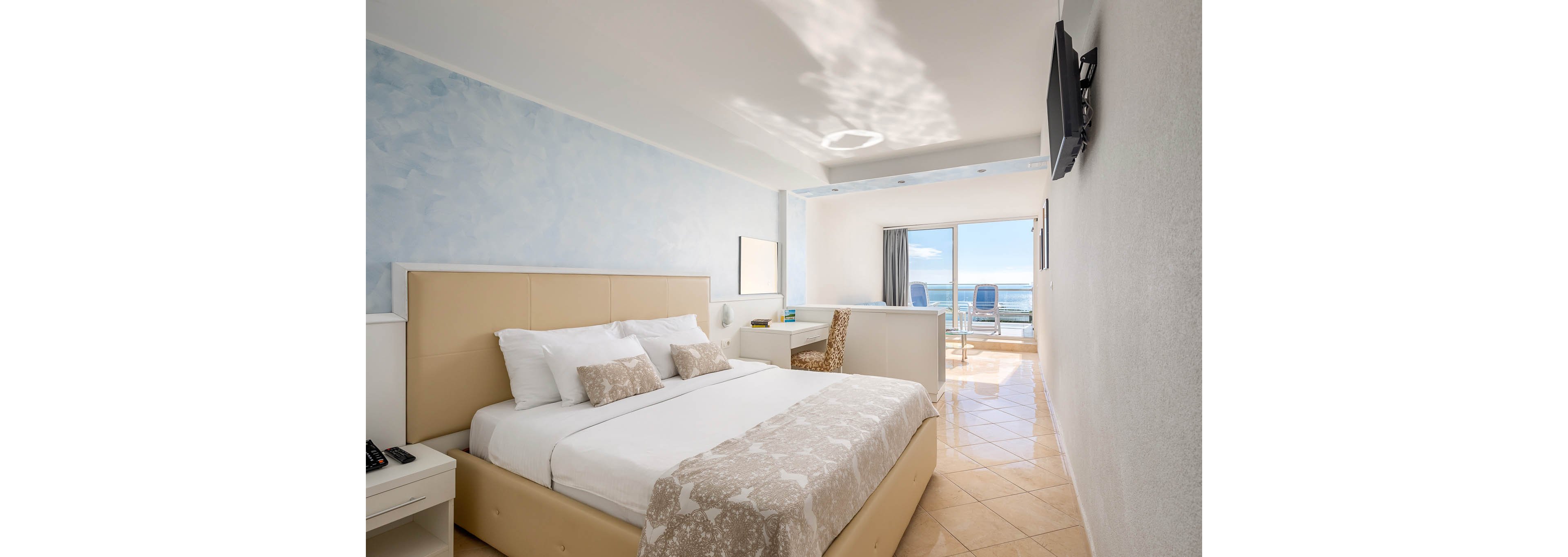 A bright, contemporary hotel room features a large bed with neutral bedding, a work desk, and a wall-mounted television. The space opens to a private balcony with two blue lounge chairs overlooking the ocean. Soft blue and beige tones create a calm, inviting atmosphere, while natural light fills the room through sliding glass doors.
