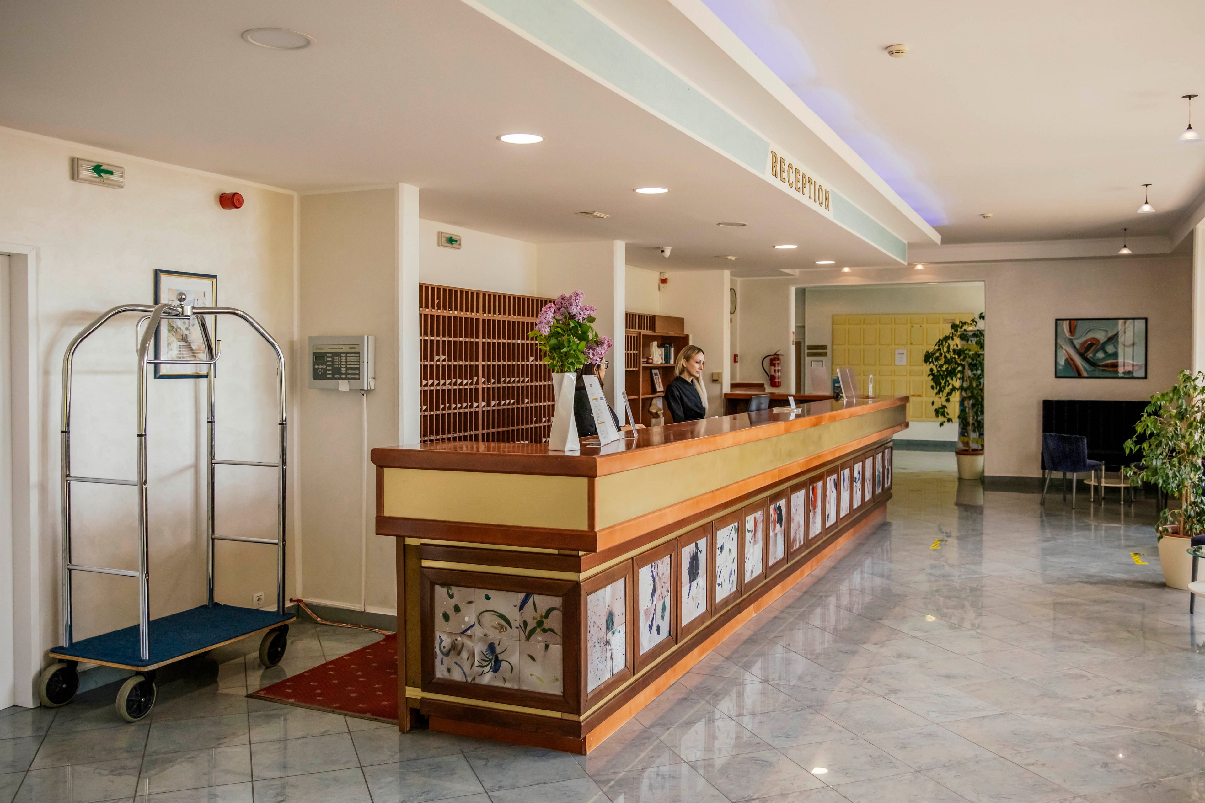 A spacious hotel reception area features a long wooden front desk with decorative panels and a concierge standing behind it. The setting is bright with polished tile floors, indoor plants, and a luggage cart visible to the side. The word 'RECEPTION' is clearly displayed above the desk, and the atmosphere is welcoming and professional. The area is well-lit with overhead lighting and has a clean, organized appearance.