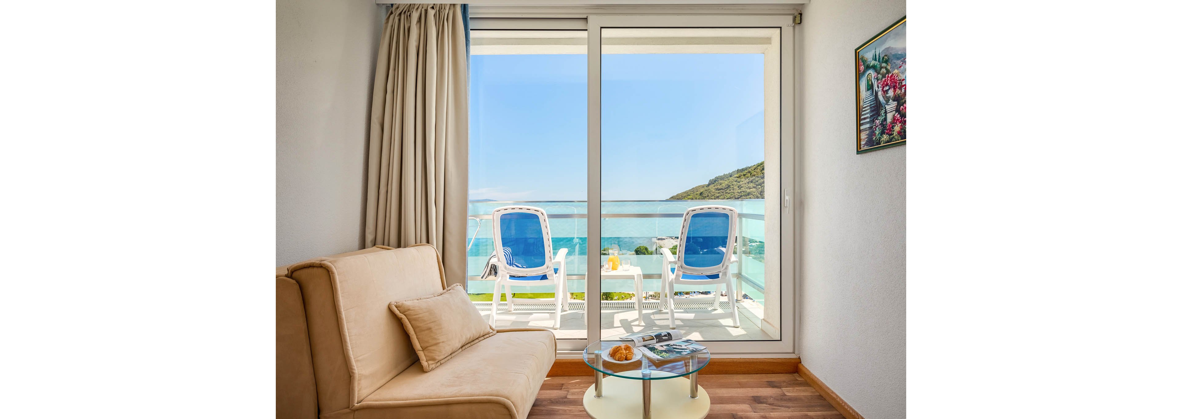 A bright, contemporary hotel room features a beige sofa and a glass coffee table with breakfast items and magazines. Through large sliding glass doors, two blue and white lounge chairs sit on a balcony overlooking a turquoise ocean and green hills. The scene is sunlit and tranquil, with a painting on the wall adding a touch of color. No visible text or numbers are present in the image.