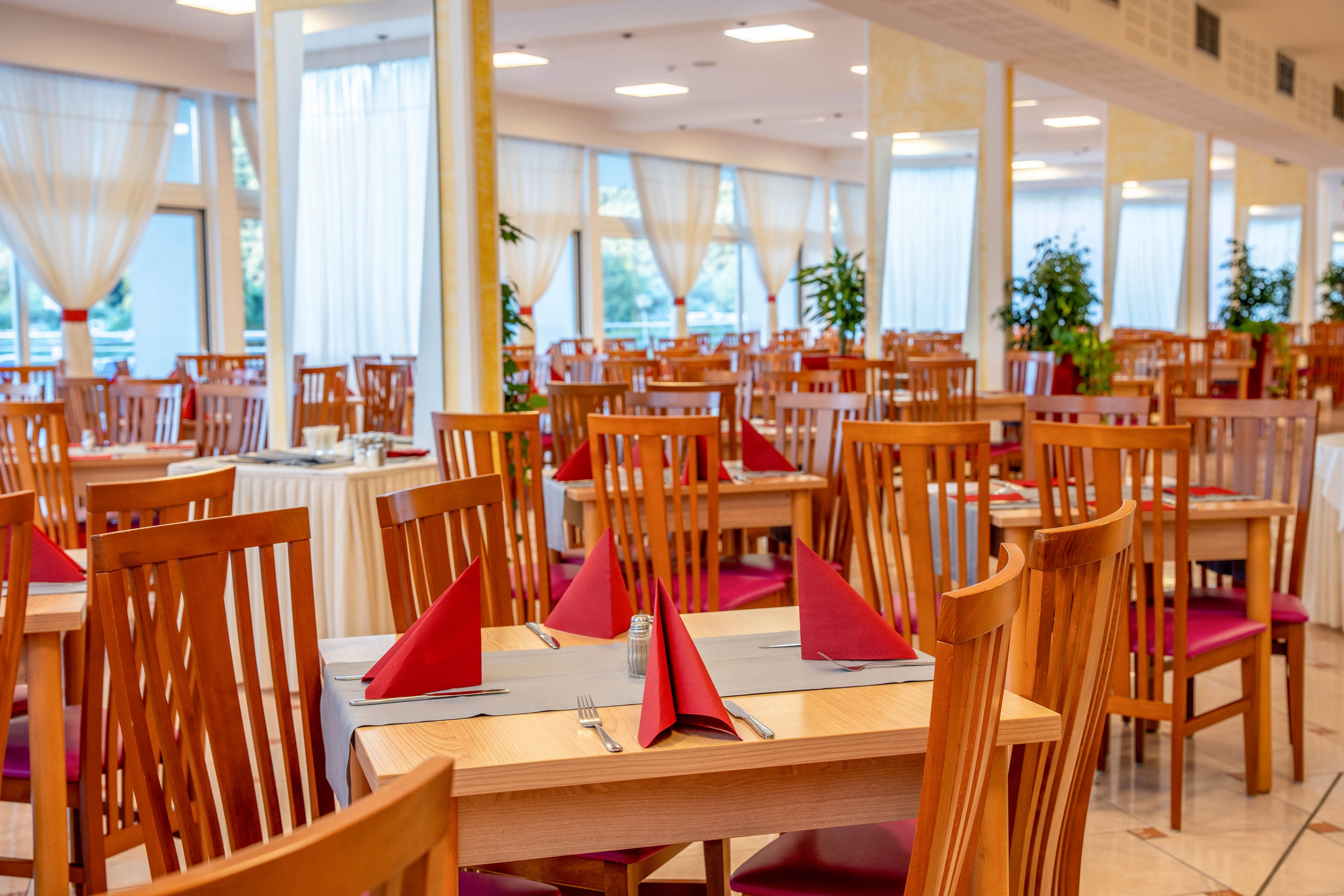 A spacious restaurant dining area features neatly arranged wooden tables and chairs, each table set with red folded napkins and place settings. Large windows with sheer white curtains allow natural light to fill the room, creating a bright and inviting atmosphere. The setting is clean and orderly, with a modern yet warm palette of wood tones and red accents. No people are present, emphasizing the serene and prepared environment.