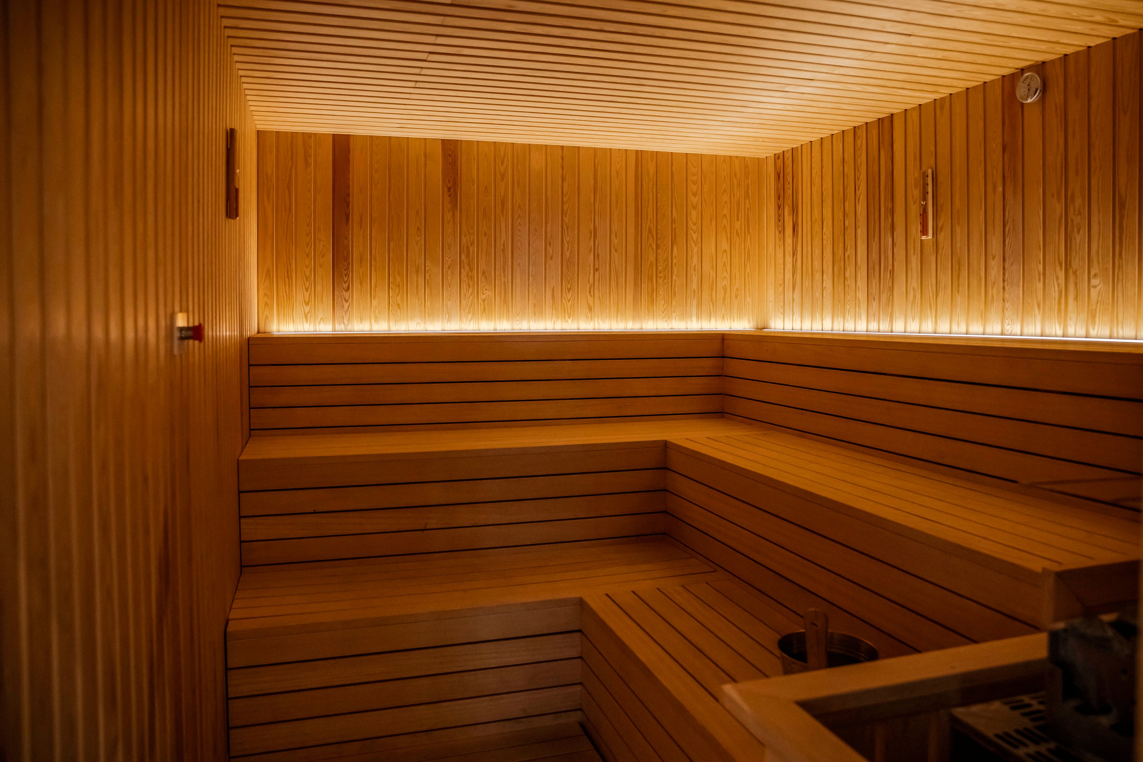 A spacious, contemporary sauna room features smooth wooden benches and walls, illuminated by soft, warm ambient lighting. The setting is empty, emphasizing the clean lines and inviting atmosphere. A small thermometer is visible on the wall, adding a functional detail to the serene environment. The overall mood is tranquil and relaxing, with a natural wood palette.