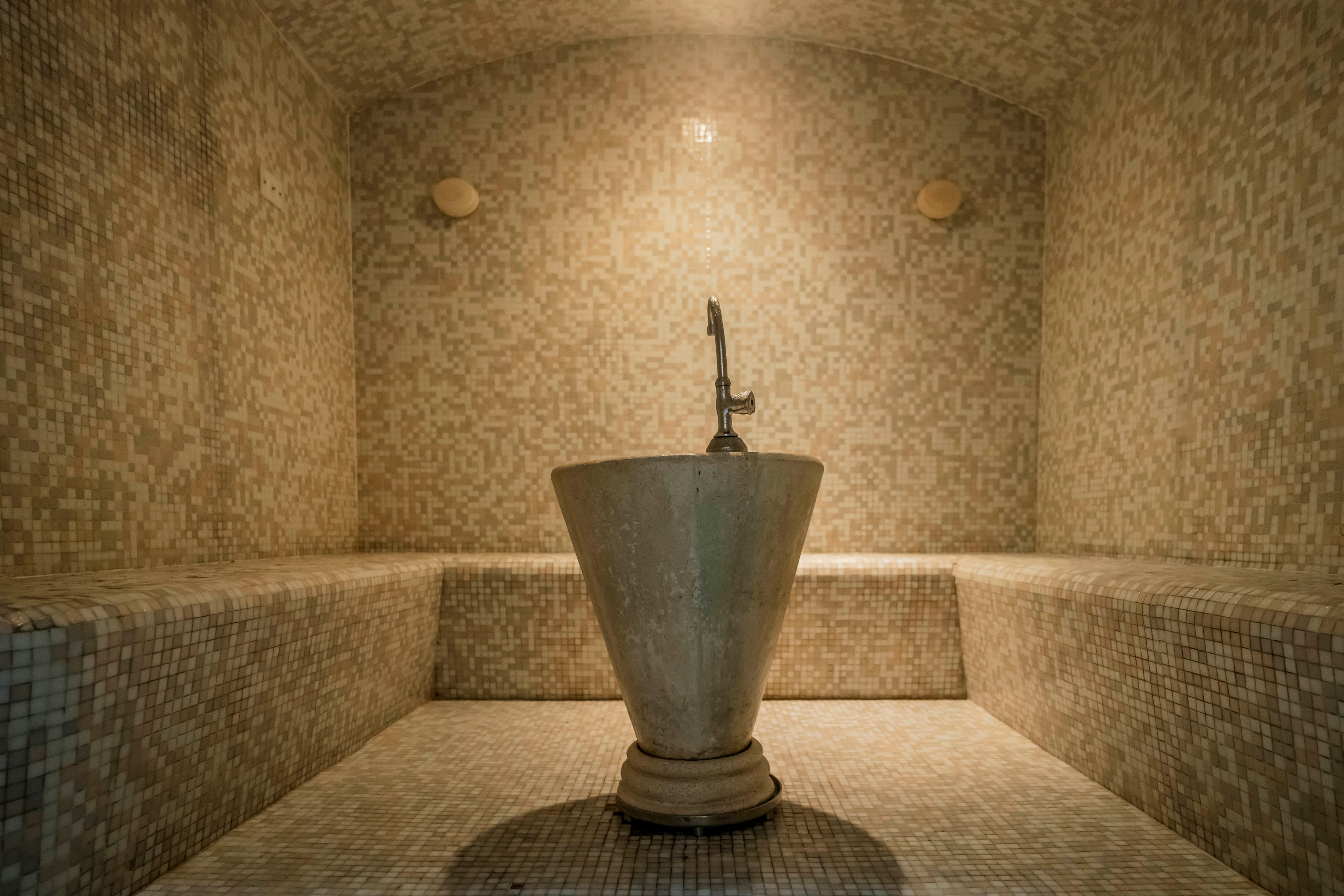 A spacious steam room features mosaic tile walls and benches, creating a serene and calming environment. At the center, a large stone basin with a faucet serves as the focal point, emphasizing the room’s traditional spa design. Warm, neutral tones dominate the palette, enhancing the tranquil and relaxing mood. The setting is empty, highlighting the architectural details and inviting atmosphere.