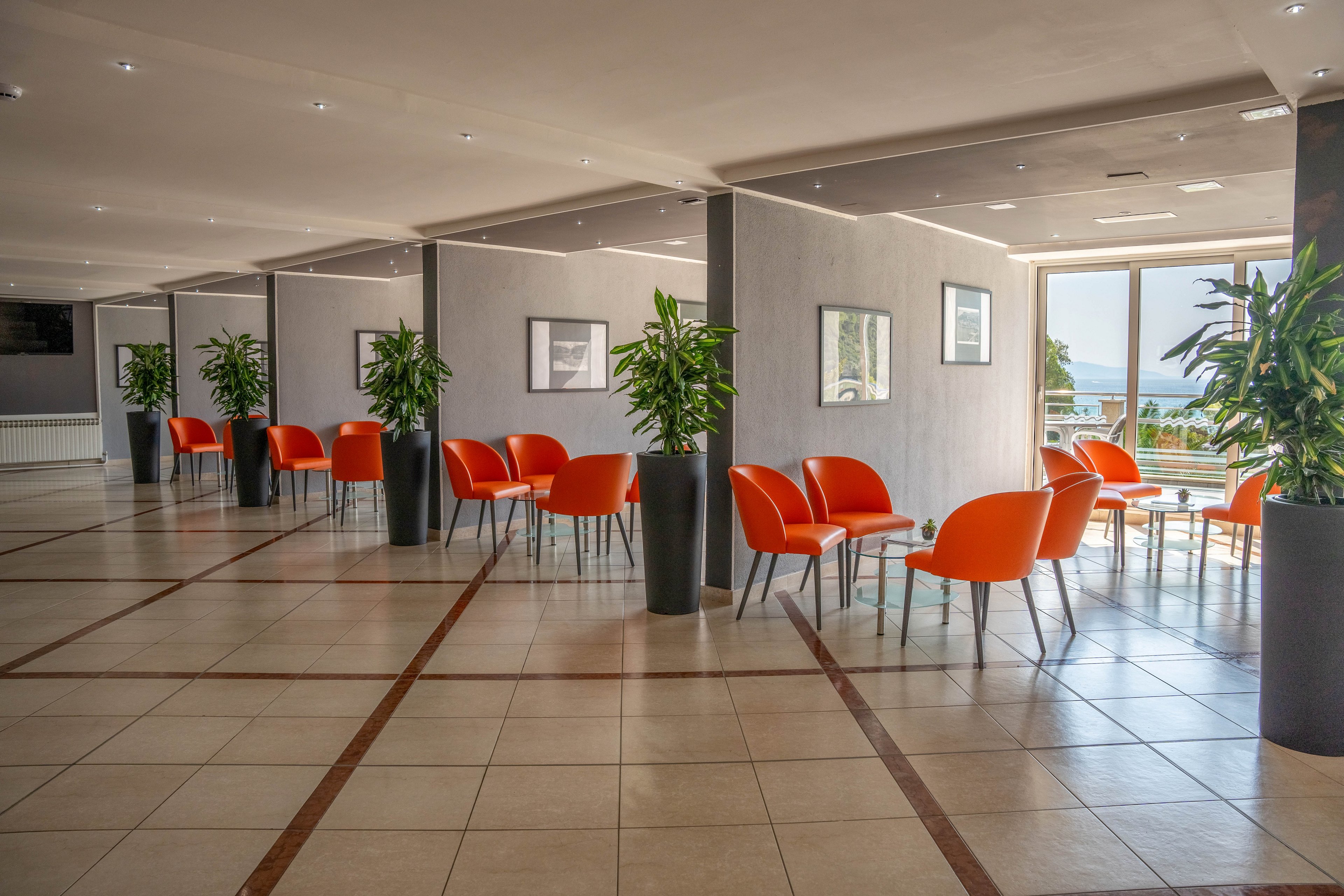A spacious, contemporary lounge area features clusters of bright orange chairs and small tables arranged along partitioned walls. Large potted plants add greenery, while framed artwork decorates the neutral-toned walls. Natural light streams in through expansive windows, creating an inviting and airy atmosphere. The tiled floor and minimalist decor contribute to a clean, modern aesthetic. No visible people or text are present in the scene.
