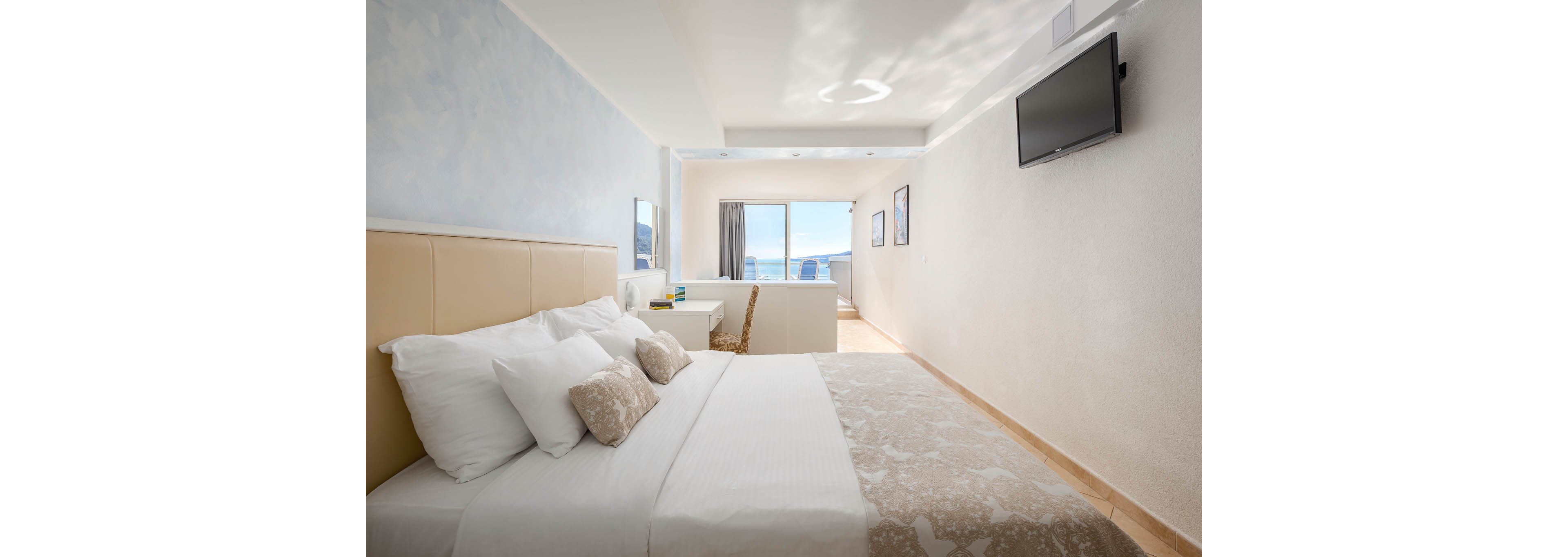 A bright, contemporary hotel room features a neatly made bed with neutral bedding and accent pillows. The space includes a wall-mounted flat-screen TV, a desk with a mirror, and large windows leading to a balcony overlooking the ocean. The room is decorated in soft beige and blue tones, creating a calm and inviting atmosphere. Natural light fills the space, highlighting the clean and minimalist design.