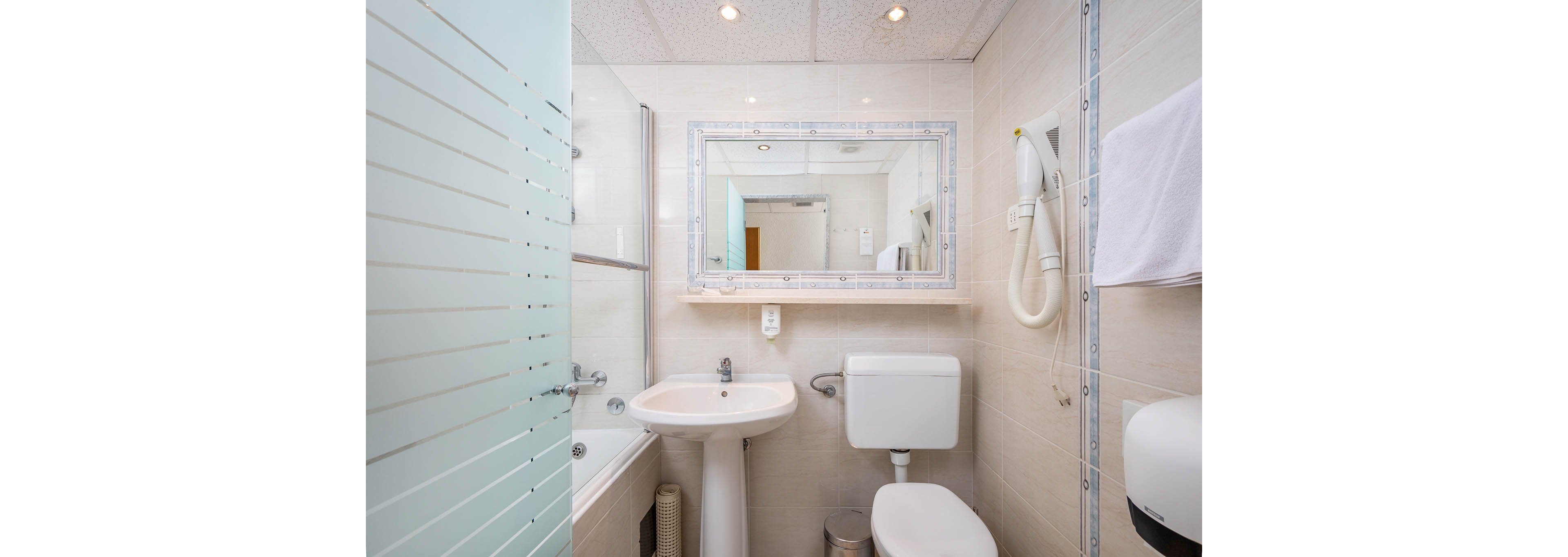 A clean, modern hotel bathroom features a large wall mirror above a white sink and toilet. The space includes a bathtub with a glass partition, a mounted hairdryer, and a towel hanging on the wall. Neutral tiles and bright lighting create a fresh, inviting atmosphere. The ceiling has recessed spotlights, and the overall palette is light and minimal.