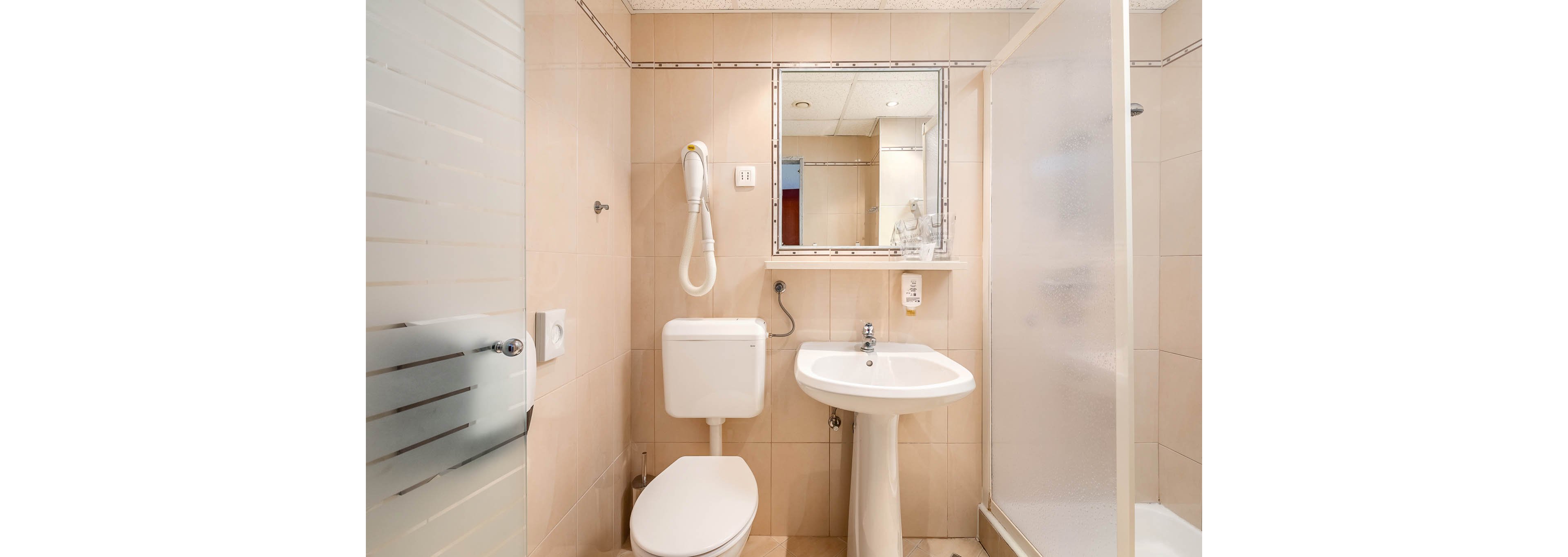 A small bathroom features a white toilet, pedestal sink, and a glass-enclosed shower. A wall-mounted hair dryer and a rectangular mirror are visible above the sink. The beige tile walls and neutral palette create a clean, simple atmosphere.