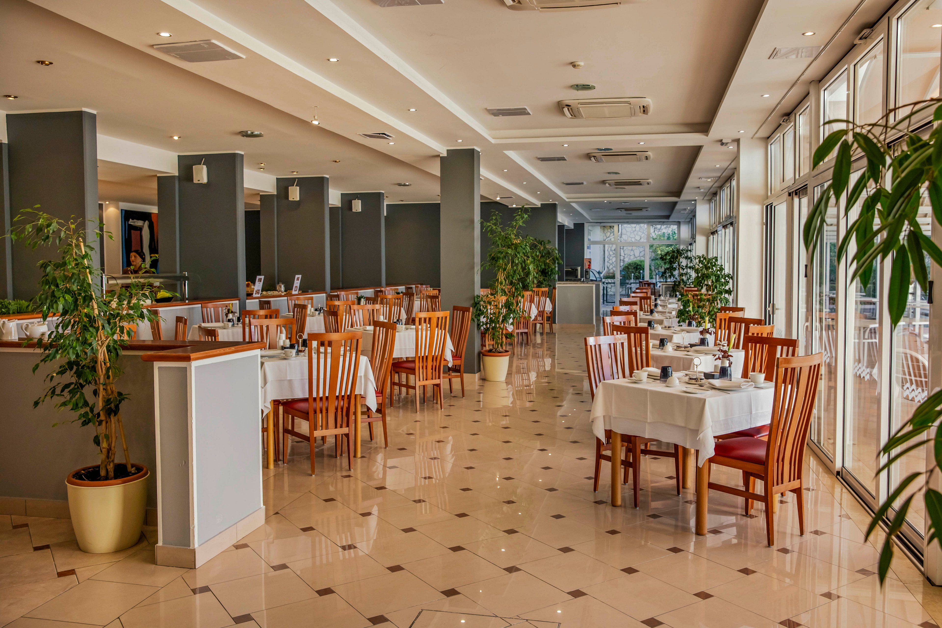 A bright, contemporary restaurant dining area features neatly arranged tables with white tablecloths and wooden chairs. Large windows allow natural light to fill the space, highlighting potted plants and a polished tile floor. The setting is clean and inviting, with a neutral color palette and minimalist decor. No people are present, emphasizing the calm and orderly atmosphere.