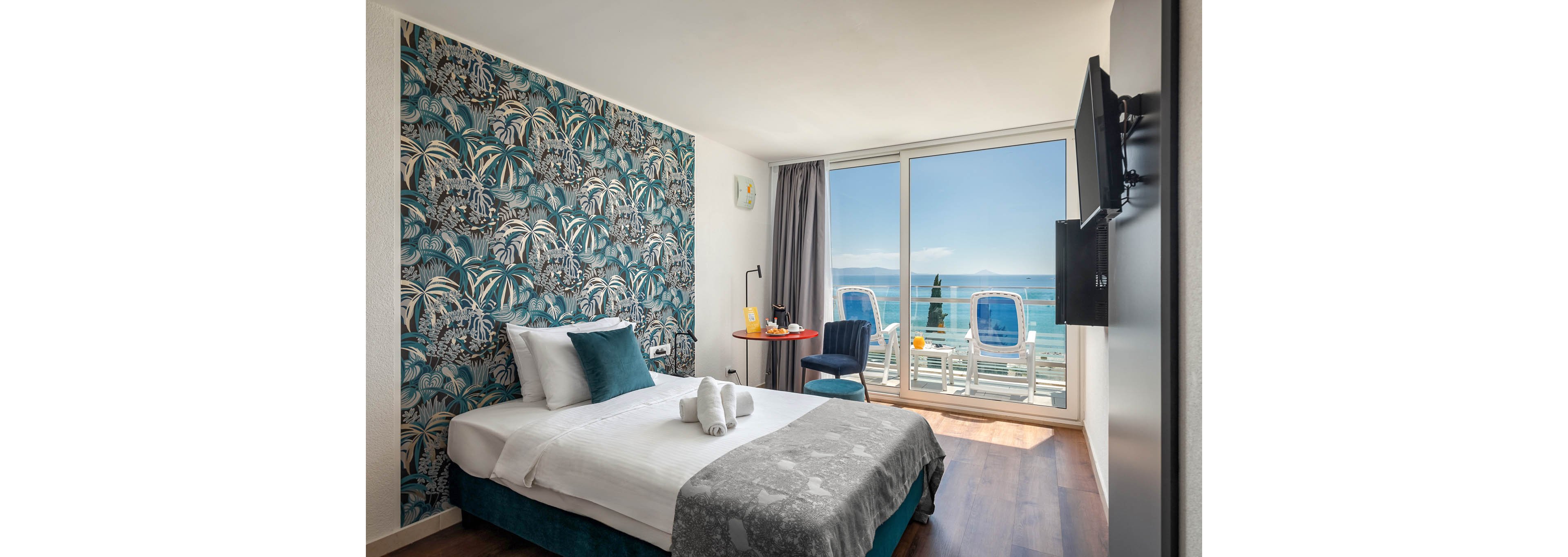 A stylish hotel room features a double bed with white linens and teal accents, set against a bold patterned accent wall. Large windows open to a balcony with lounge chairs overlooking a bright blue ocean. The room includes a small desk, chair, and a wall-mounted television, with sunlight streaming in to create a fresh, inviting atmosphere. No visible text or numbers are present in the image.