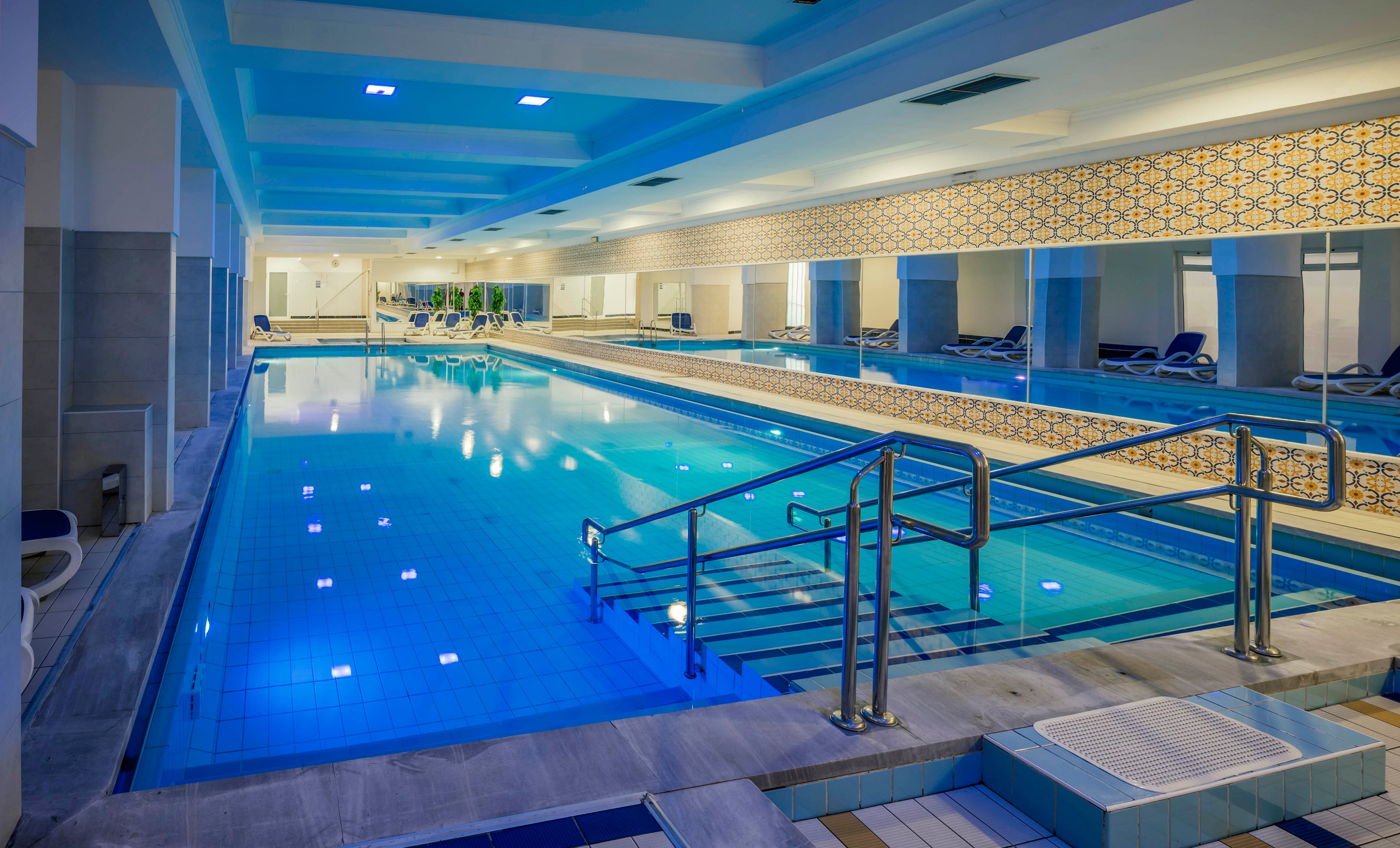 A spacious indoor swimming pool is featured in a contemporary facility with clean lines and blue-tinted lighting. The pool area includes metal handrails, tiled flooring, and several lounge chairs along the sides. The walls are decorated with geometric patterns, and the atmosphere is calm and inviting. No people are present, emphasizing the serene and relaxing environment.