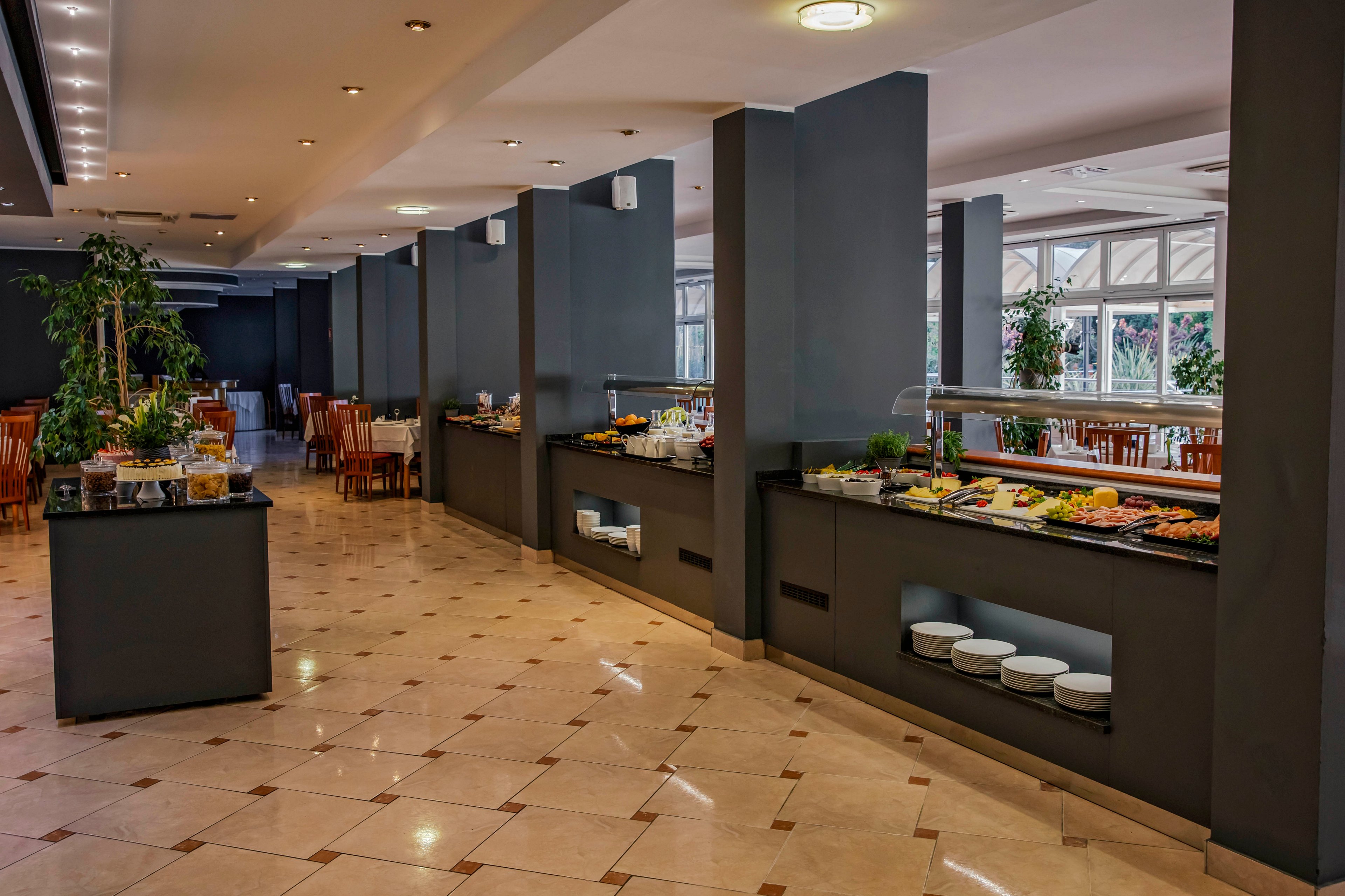 A spacious hotel buffet area features a variety of food items arranged on long counters. The setting is clean and modern, with dark walls and tiled floors, and natural light streaming in from large windows. Empty tables and chairs are visible in the background, suggesting a calm, early or off-peak dining time. The overall mood is inviting and organized, with a neutral color palette and contemporary style.