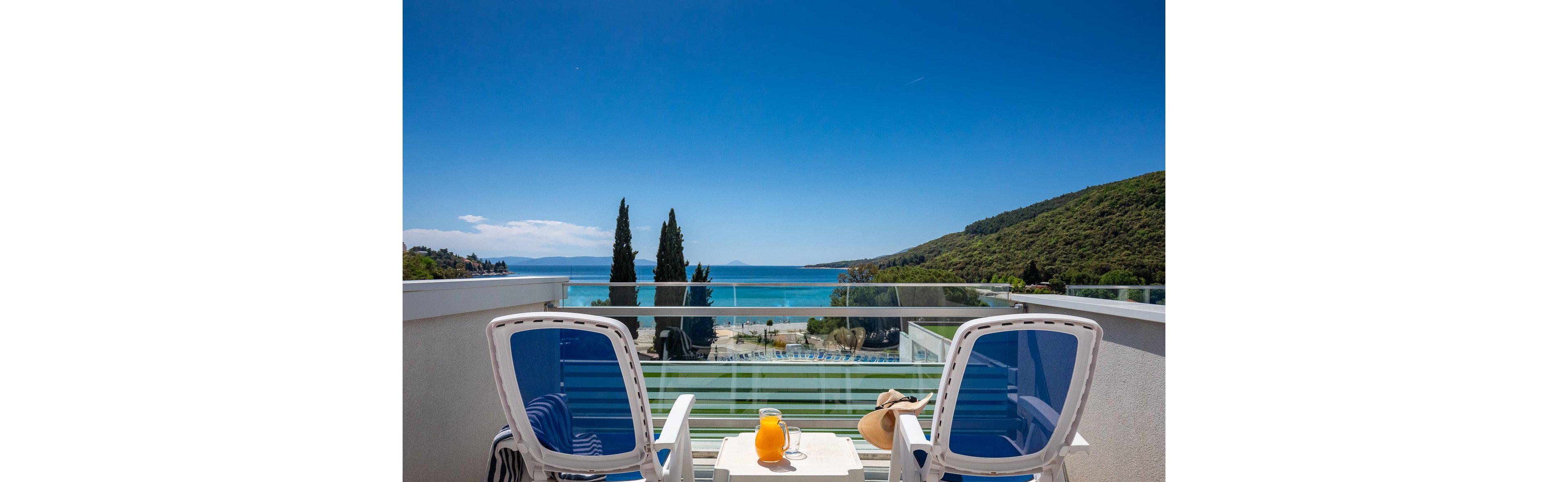 Two white chairs face a small table with a pitcher of orange juice and glasses on a balcony. The view overlooks a sandy beach, turquoise sea, and lush green hills under a clear blue sky. A sunhat and sunglasses rest on one chair, suggesting a relaxed vacation mood. Tall cypress trees and modern glass railing add to the scenic, tranquil setting.