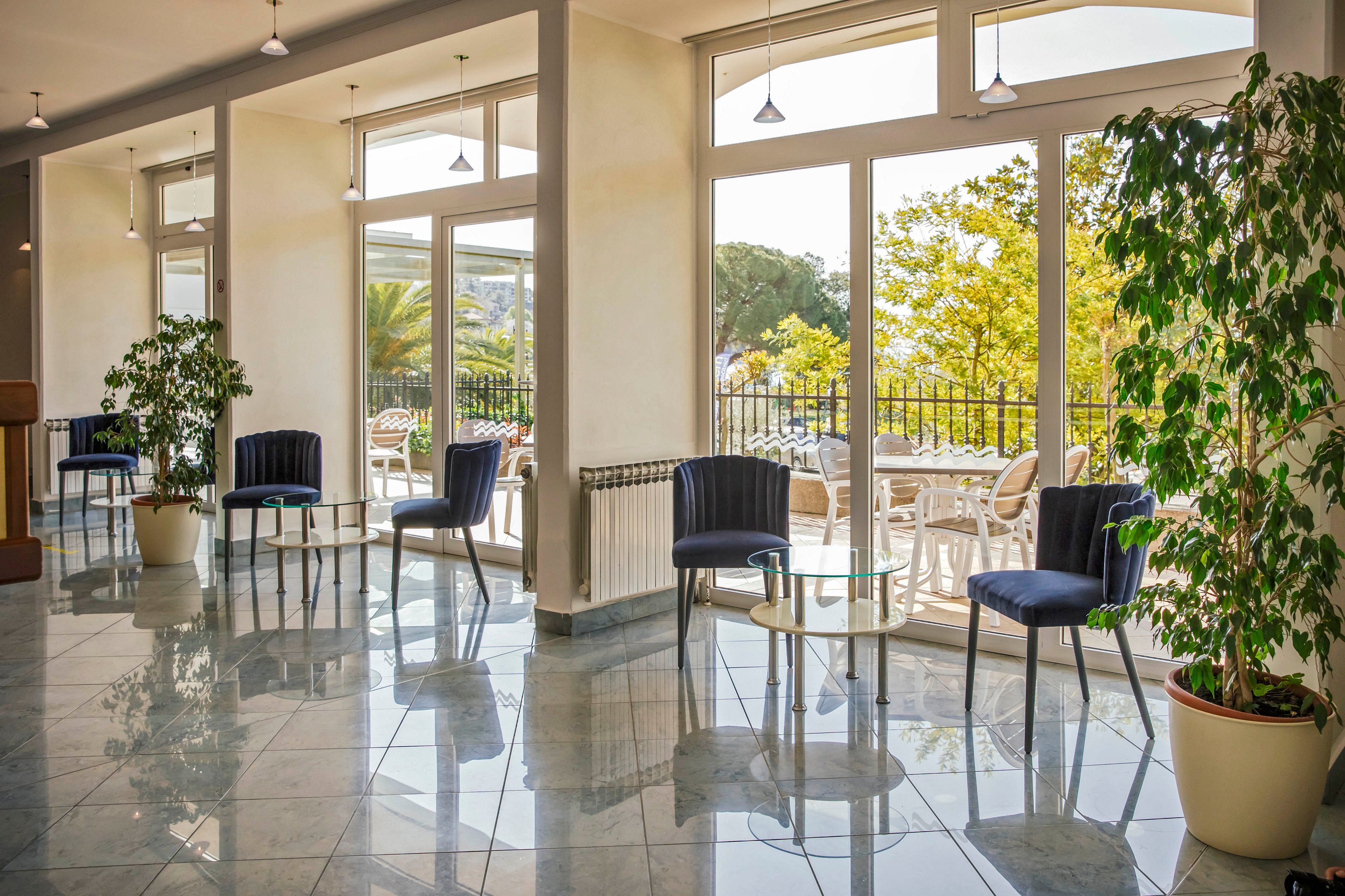 A spacious lounge area features several dark blue chairs and small round tables arranged near large windows. Sunlight streams in, illuminating the glossy tiled floor and lush indoor potted plants. Outside, a terrace with white chairs and greenery is visible, creating a relaxed and inviting atmosphere. The setting is quiet, with no people present, and the overall mood is airy and tranquil.