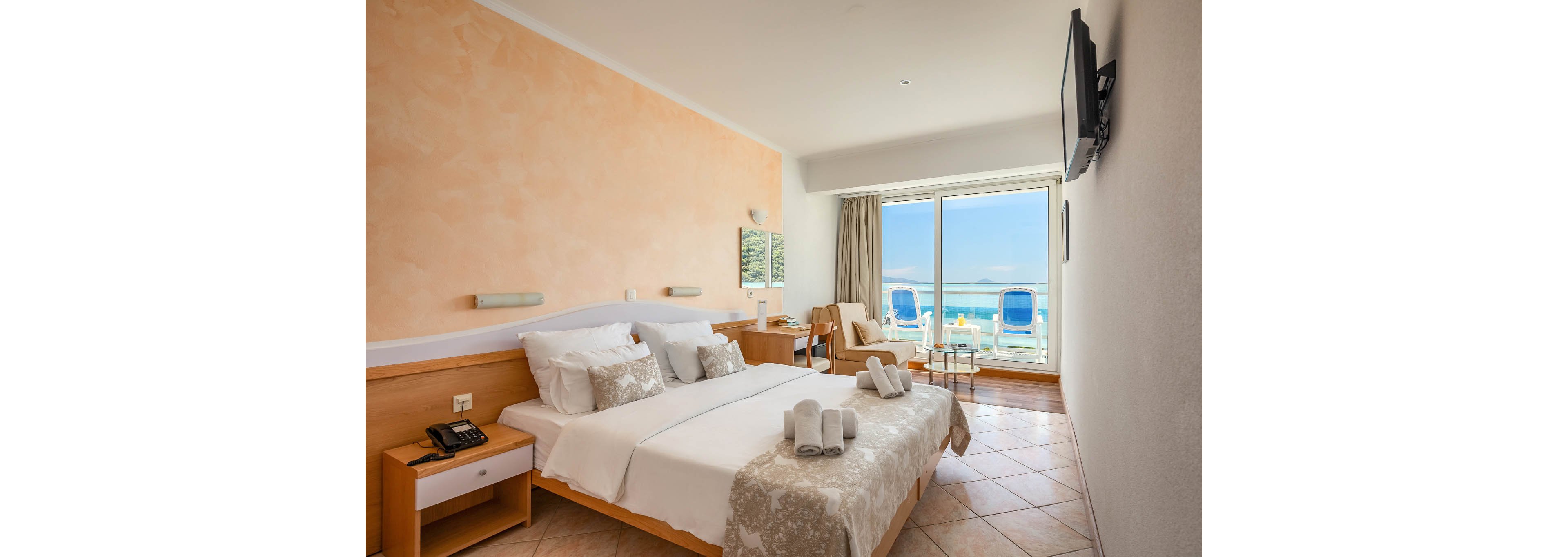 A bright, contemporary hotel room features a double bed with decorative pillows and towels, a wall-mounted television, and a small seating area. Large glass doors open to a balcony with two blue lounge chairs overlooking the ocean. The room is styled in neutral tones with tiled flooring and light wood furniture, creating a relaxing and inviting atmosphere.