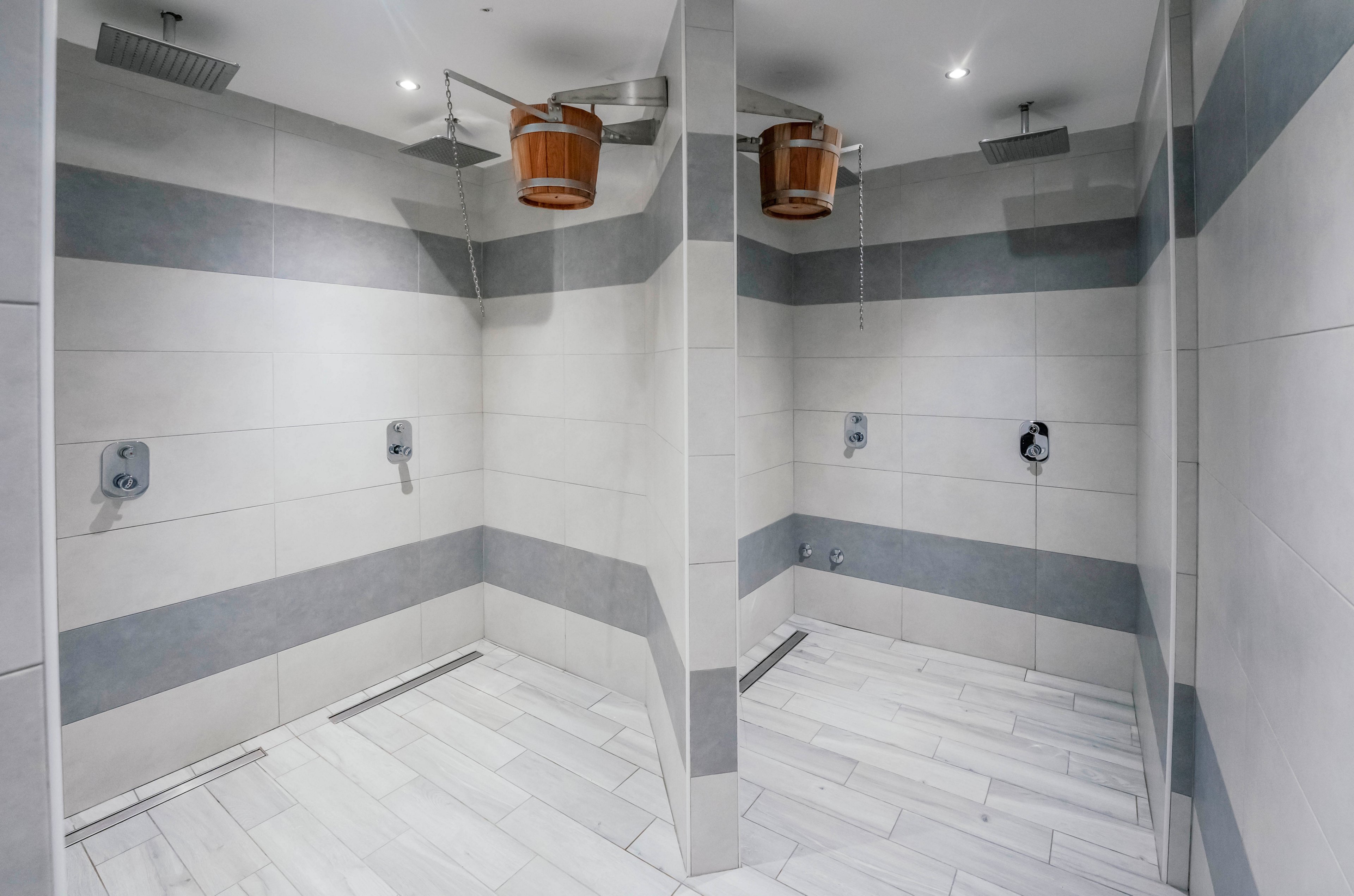 Two adjacent shower stalls feature contemporary tile walls and floors, each equipped with a wooden water bucket and overhead showerhead. The setting appears to be a spa or wellness center, with a clean, minimalist design and soft lighting. The space is unoccupied, emphasizing the sleek lines and neutral color palette. No visible text or numbers are present in the image.