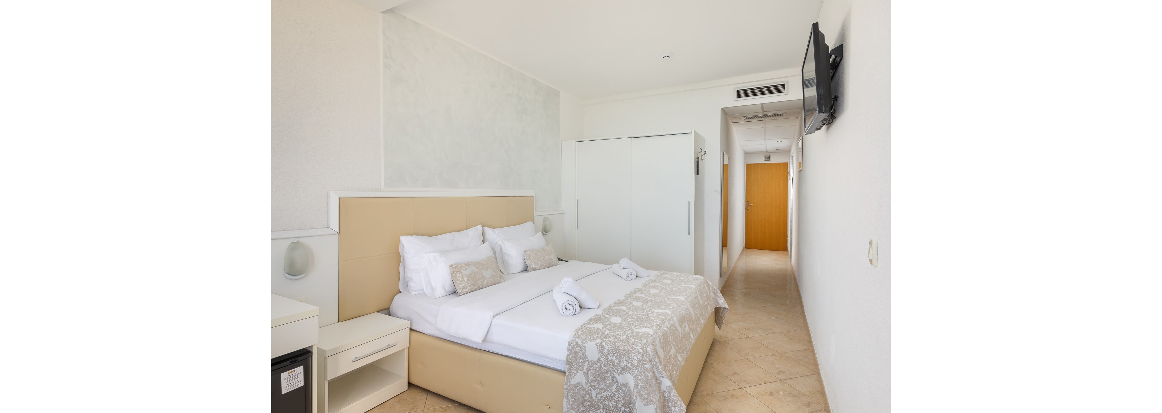 A bright, modern hotel room features a neatly made double bed with white linens and decorative pillows. The room includes a wall-mounted television, a wardrobe, and a small refrigerator. Neutral tones and minimal decor create a clean, inviting atmosphere. The hallway leads to a wooden door, and the overall palette is light and airy.