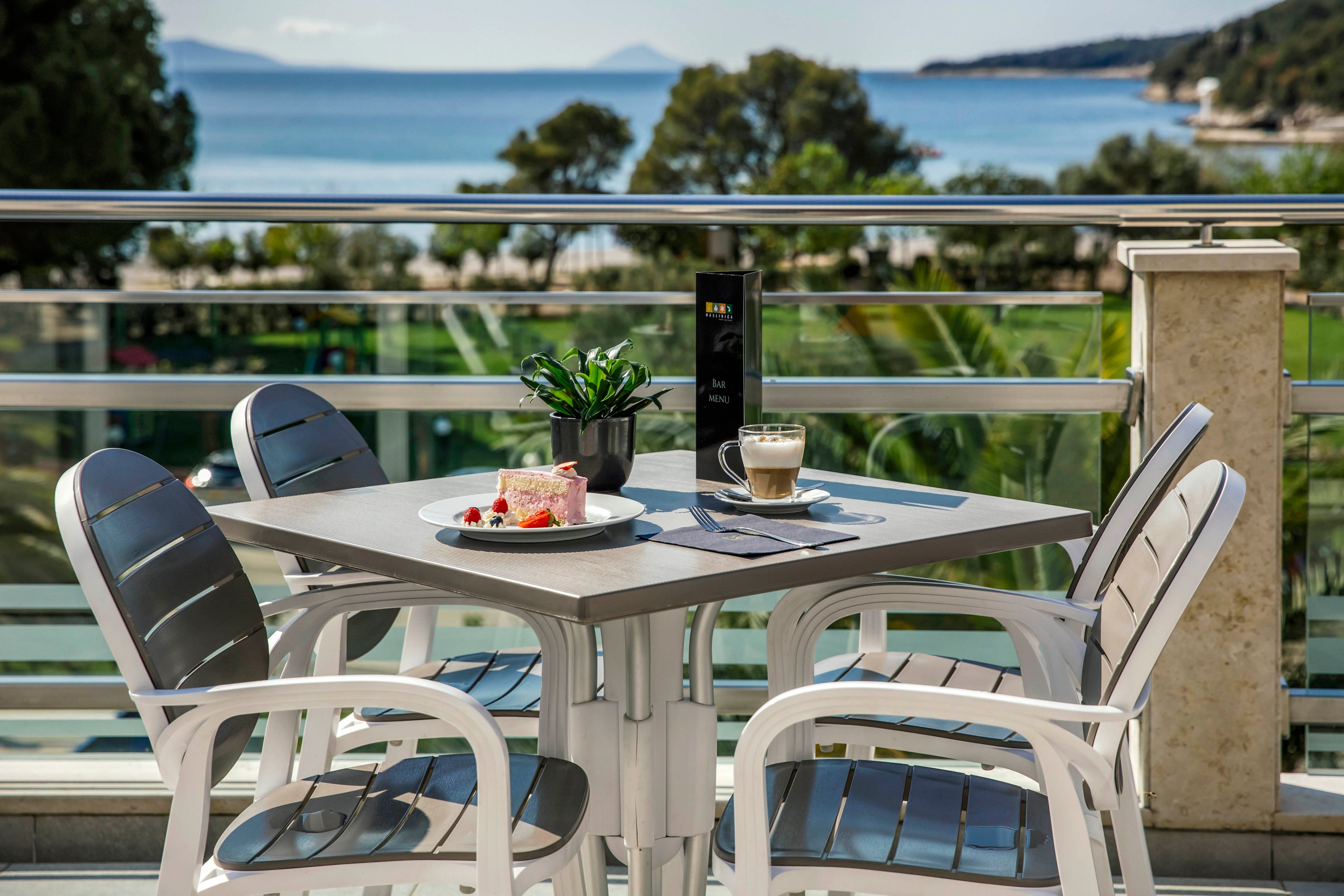 A modern outdoor café table is set with a slice of cake, a cup of coffee, and a small potted plant. The scene overlooks a scenic seaside view with blue water, distant mountains, and lush greenery. Four empty chairs surround the table, suggesting a tranquil and inviting atmosphere. The setting is bright and airy, with natural daylight and a relaxed mood. No visible text or numbers are present in the image.
