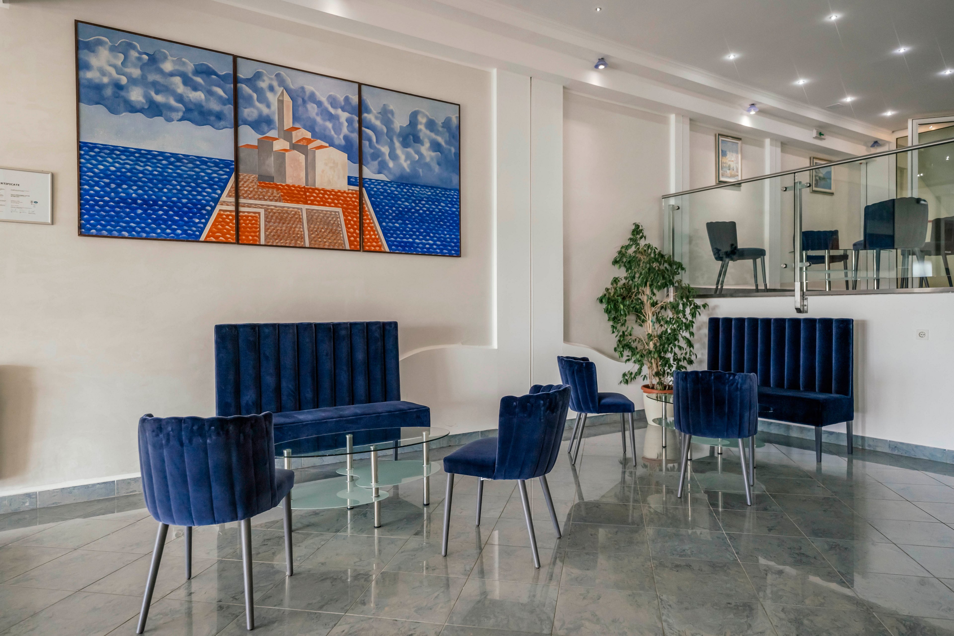 A contemporary hotel lobby features blue velvet chairs and benches arranged around glass tables. The setting is bright and clean, with a large colorful painting of a seaside town on the wall. The space includes marble flooring, a potted plant, and a glass railing on the upper level. The overall mood is calm and inviting, with a minimalist and modern style.